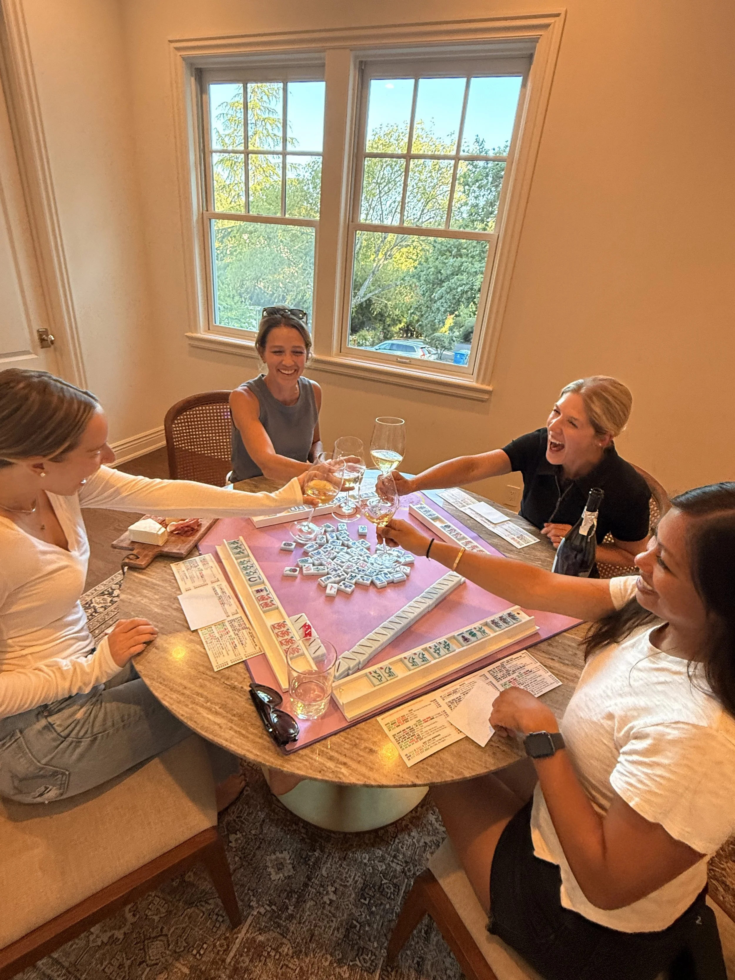 Four women playing American Mahjong, and celebrating with drinks around a wooden table in a room with large windows showing trees outside.
