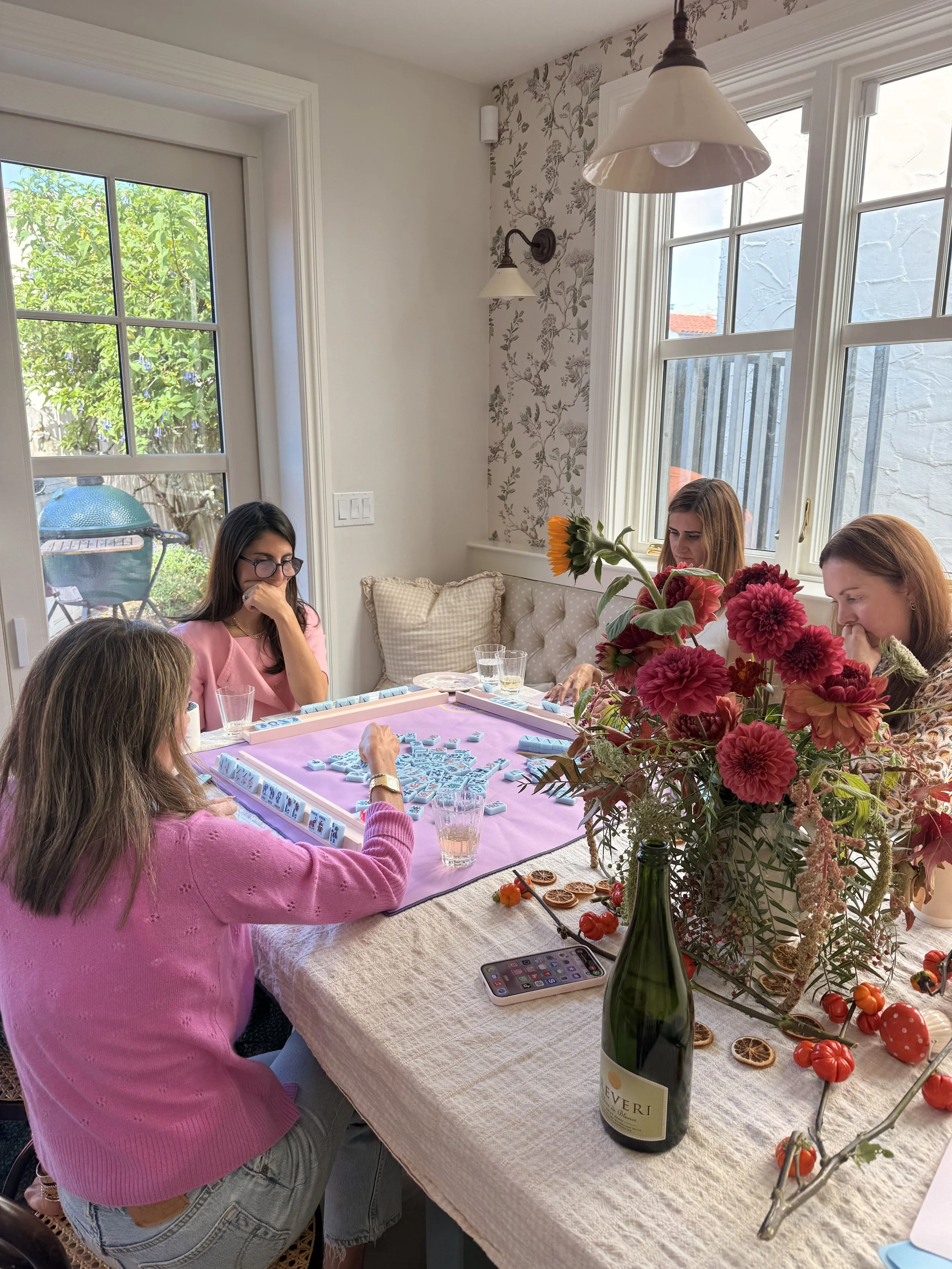 Four women playing a game of Mahjong at a dining table decorated with a vase of flowers, a bottle of wine, and some snacks.