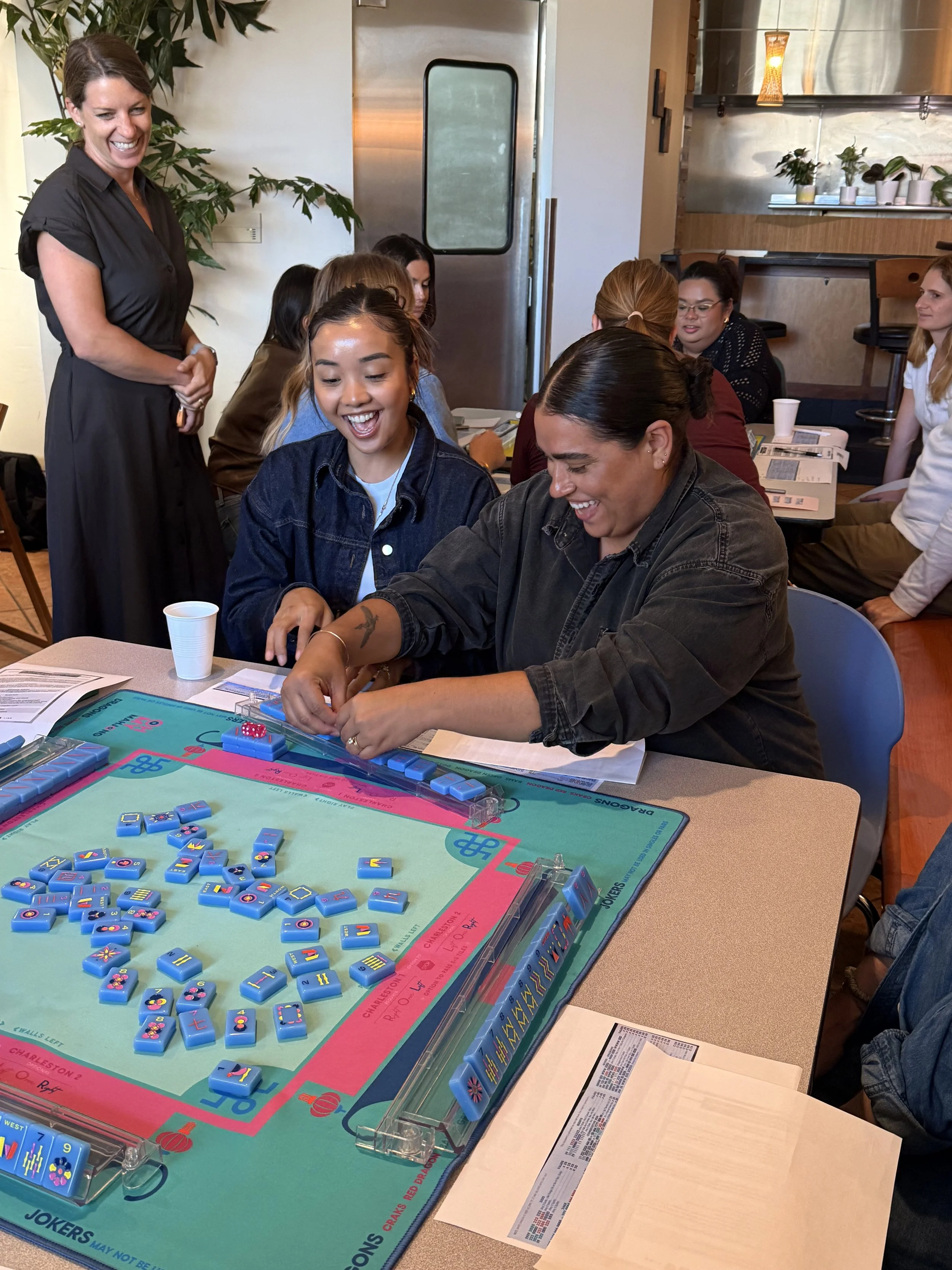 Group of women playing a game of Mahjong at a table, laughing and smiling, with a woman standing nearby.