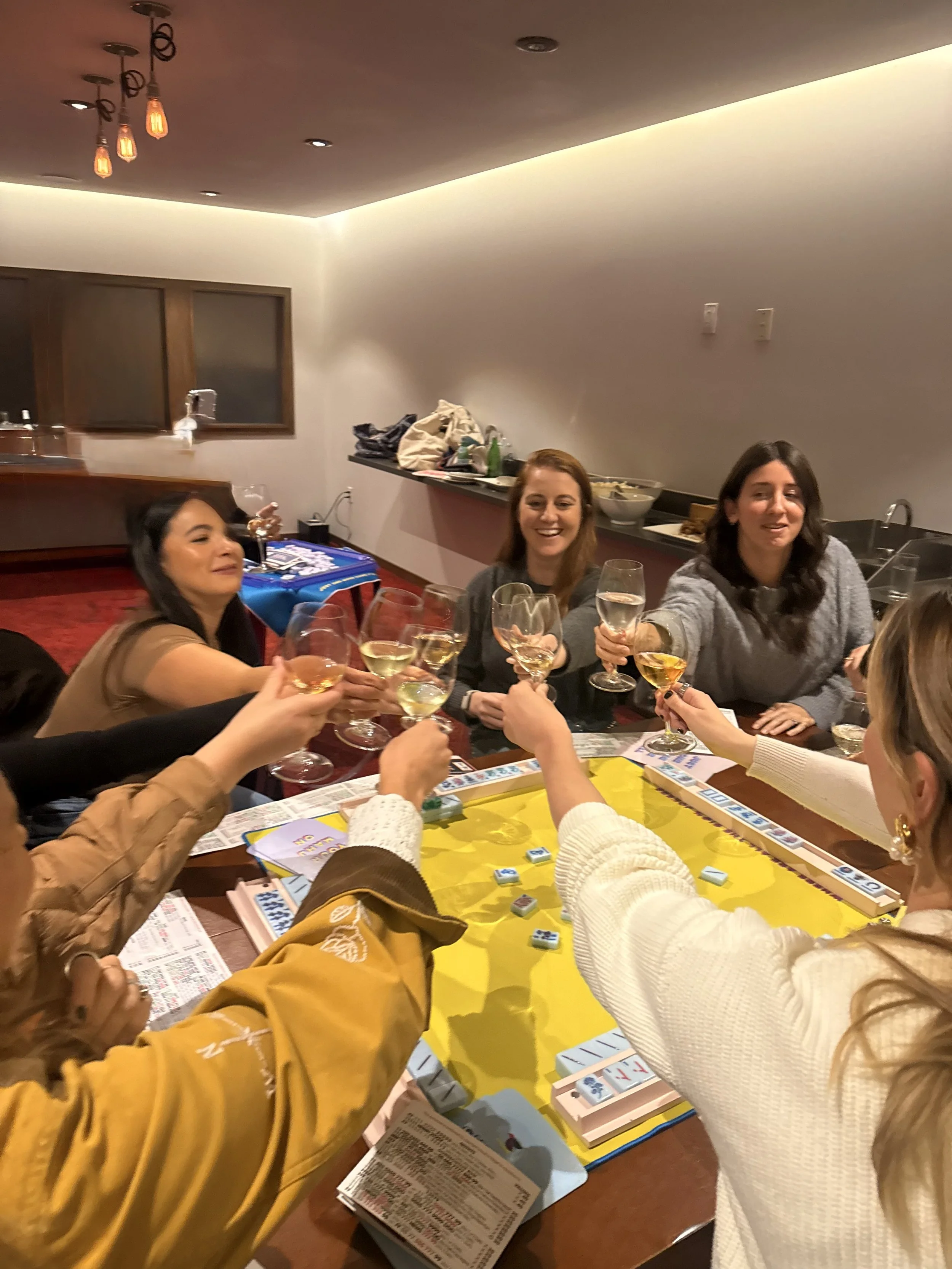 Group of women playing American Mahjong, cheers to the bird bam, with wine glasses in a cozy indoor setting.
