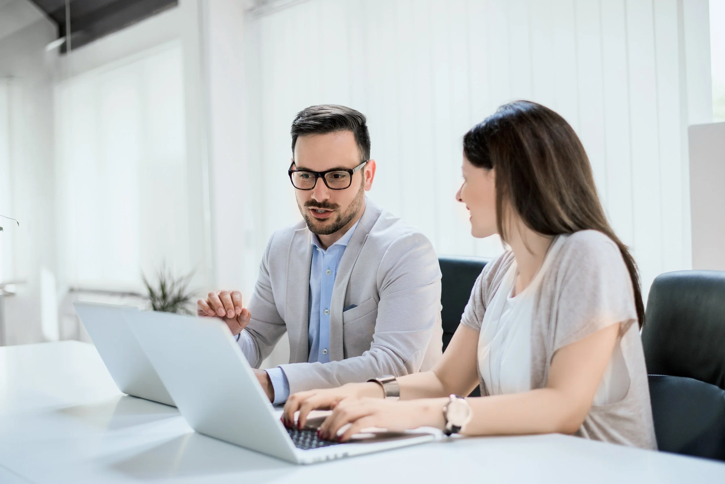 A man and woman sit at a white table in an office, looking at a laptop. The man has dark hair, glasses, and a light gray blazer, while the woman has brown hair and is wearing a light-colored blouse and cardigan.