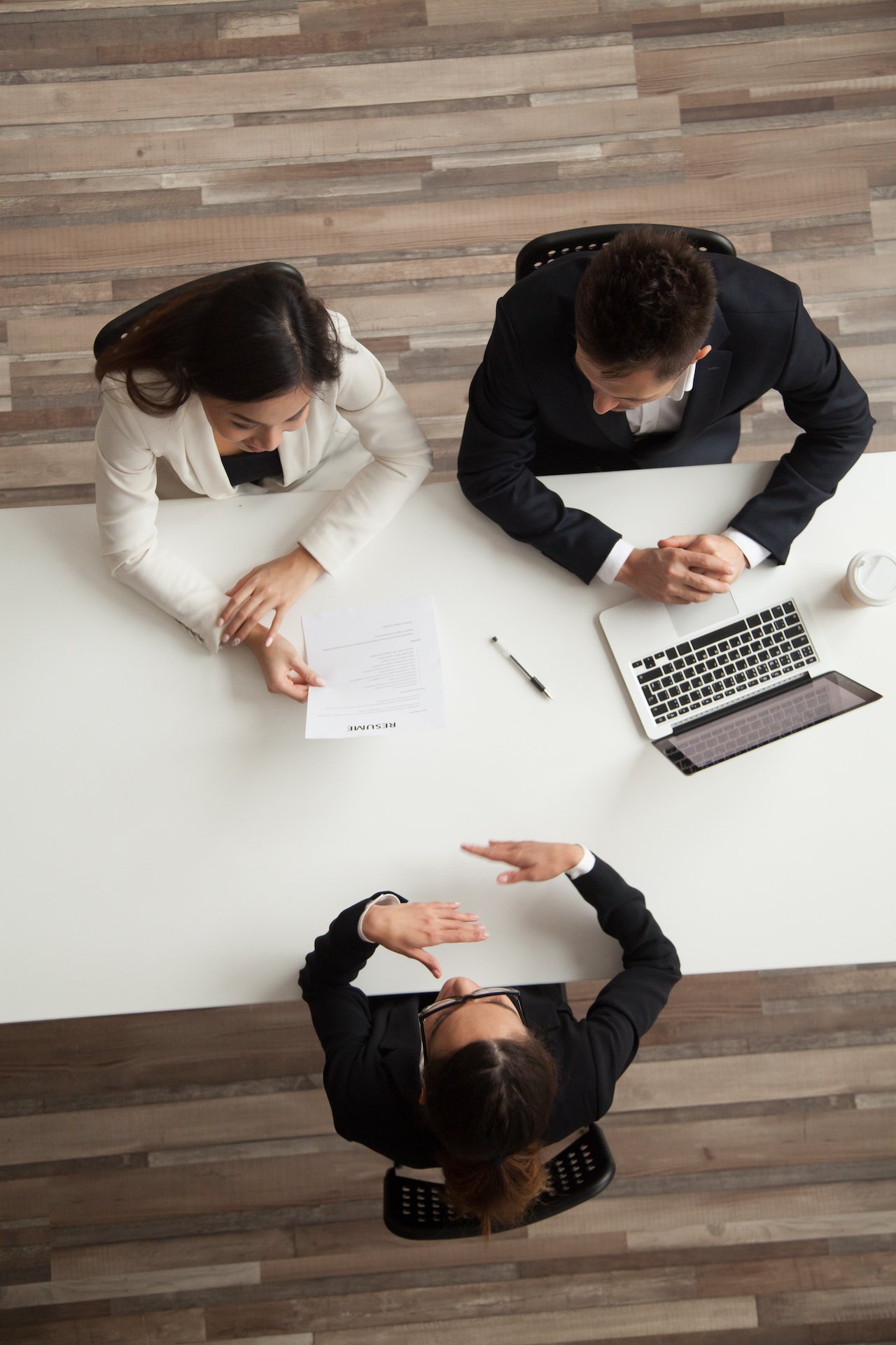 Three professionals having a discussion at a white conference table, with a woman holding a resume, a man with a laptop, and another woman gesturing during a meeting.