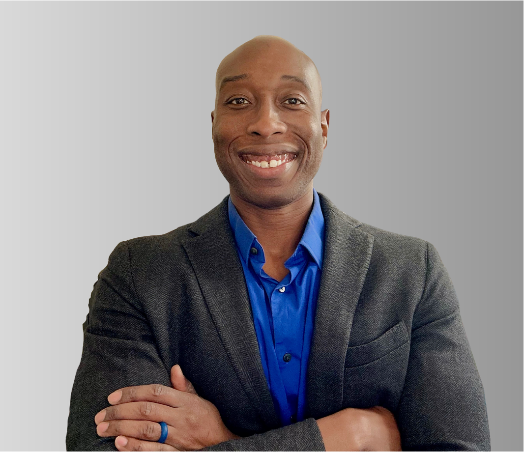 A smiling man in a blue dress shirt and gray blazer standing with arms crossed against a plain background.