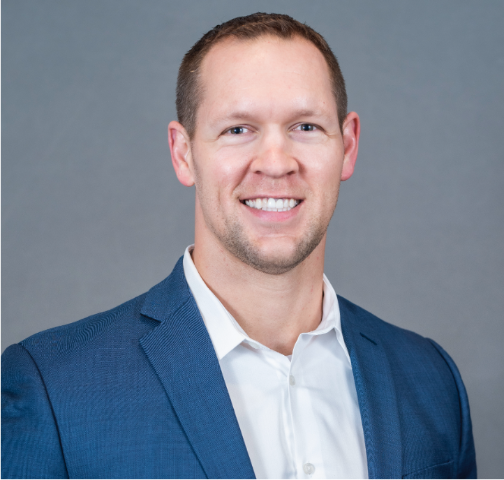 Headshot of a man in a blue suit and white shirt smiling against a gray background.