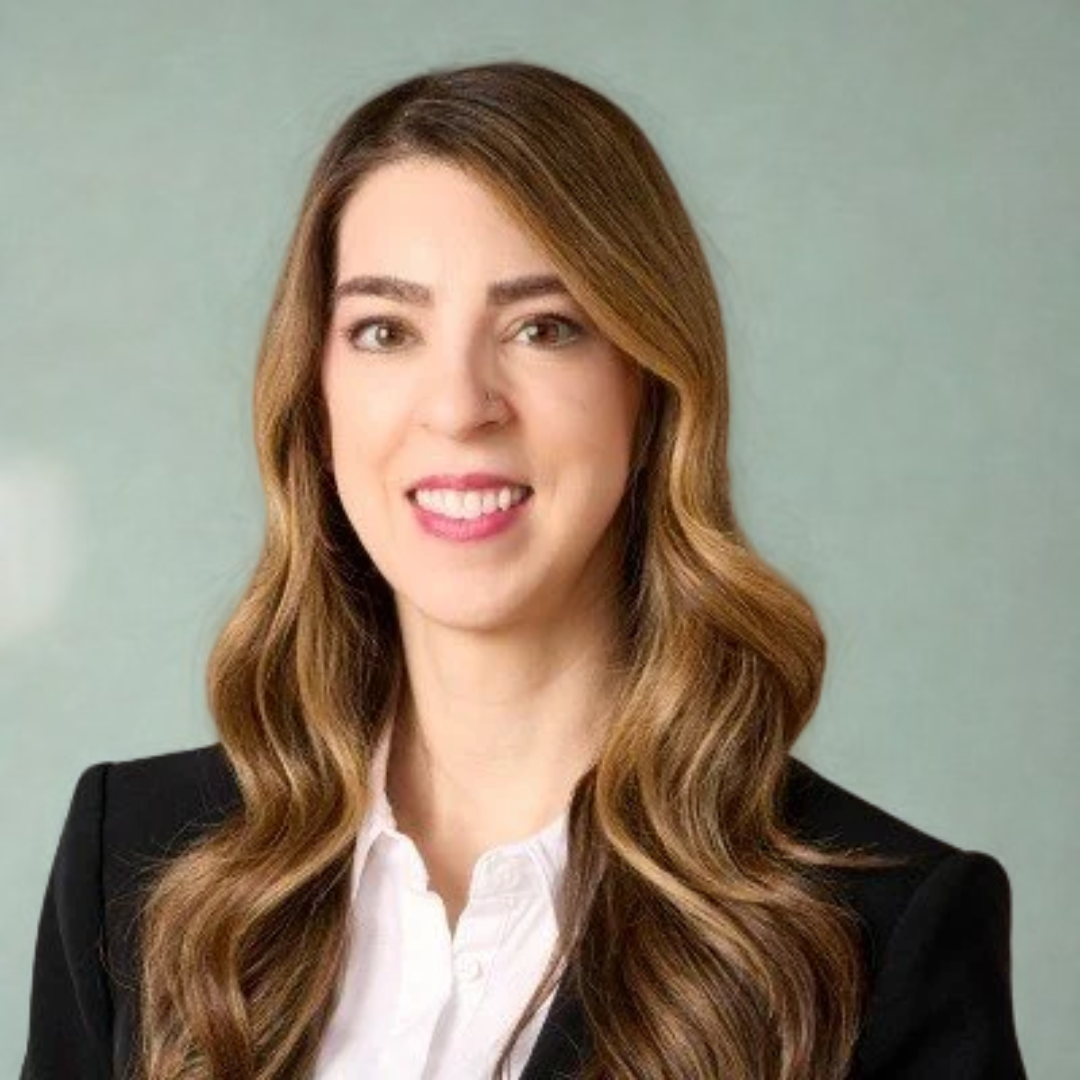 Professional woman with long wavy hair wearing a black blazer and white shirt, smiling in front of a cityscape background.