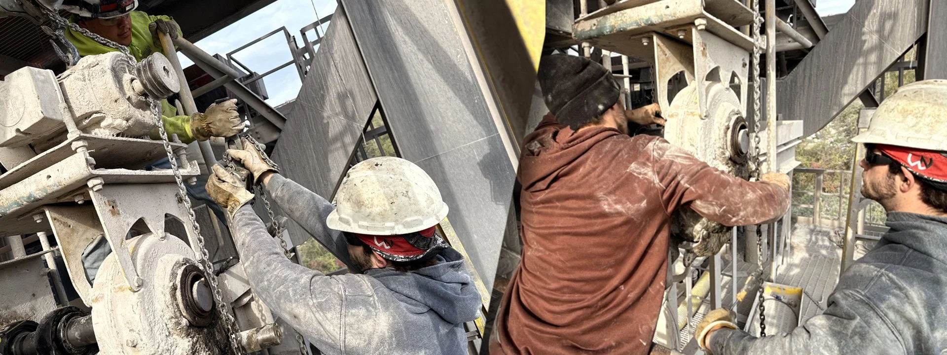 Two workers wearing safety gear, including helmets and gloves, are repairing or maintaining large industrial machinery outdoors. They are using tools and working closely together on the machinery with a metal framework and chains around.