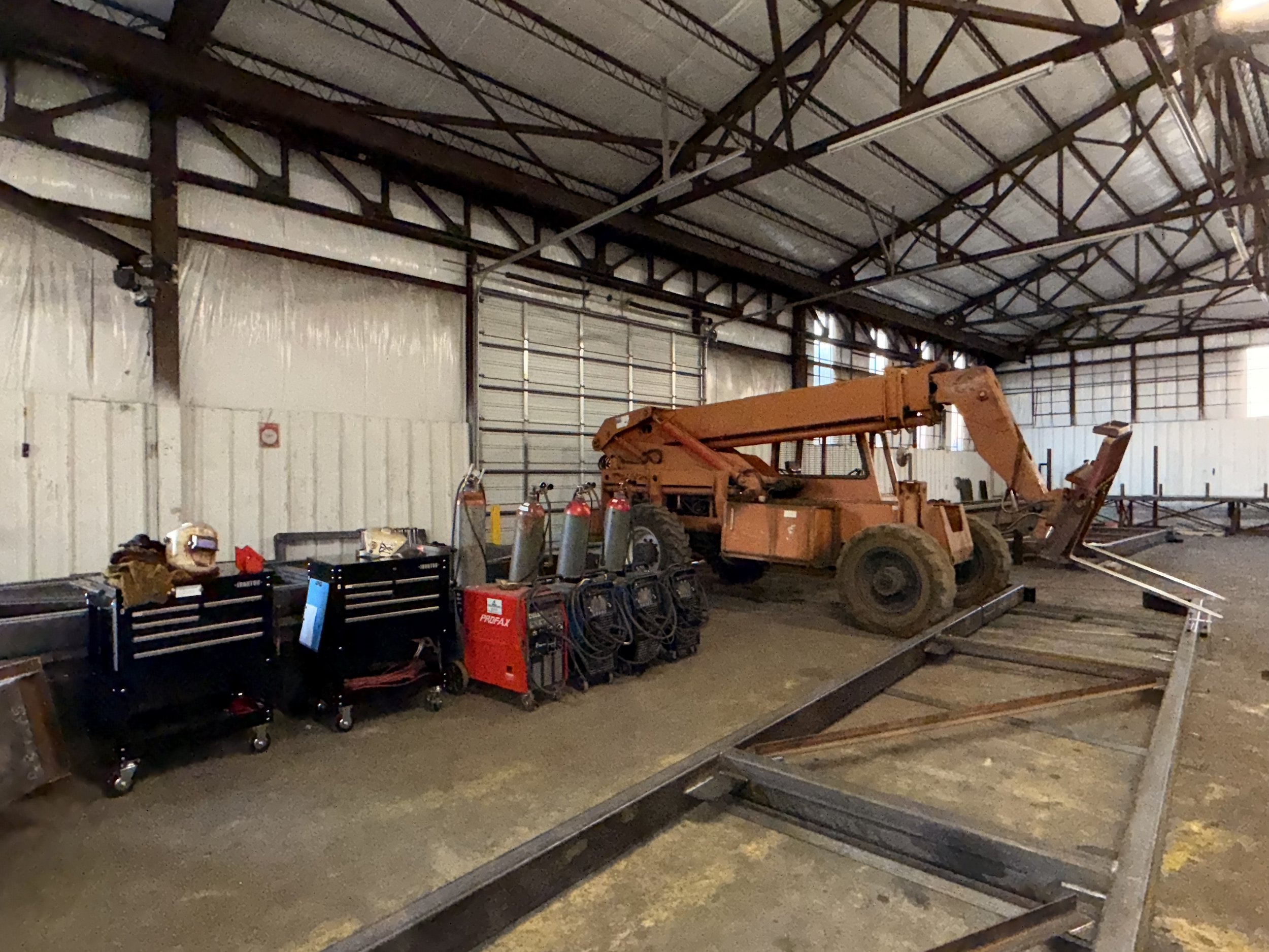 An orange telescopic boom lift inside a warehouse or garage with tools and equipment on the left side, including three welding machines and oxygen tanks, and a partially disassembled metal frame or track on the floor in the foreground.
