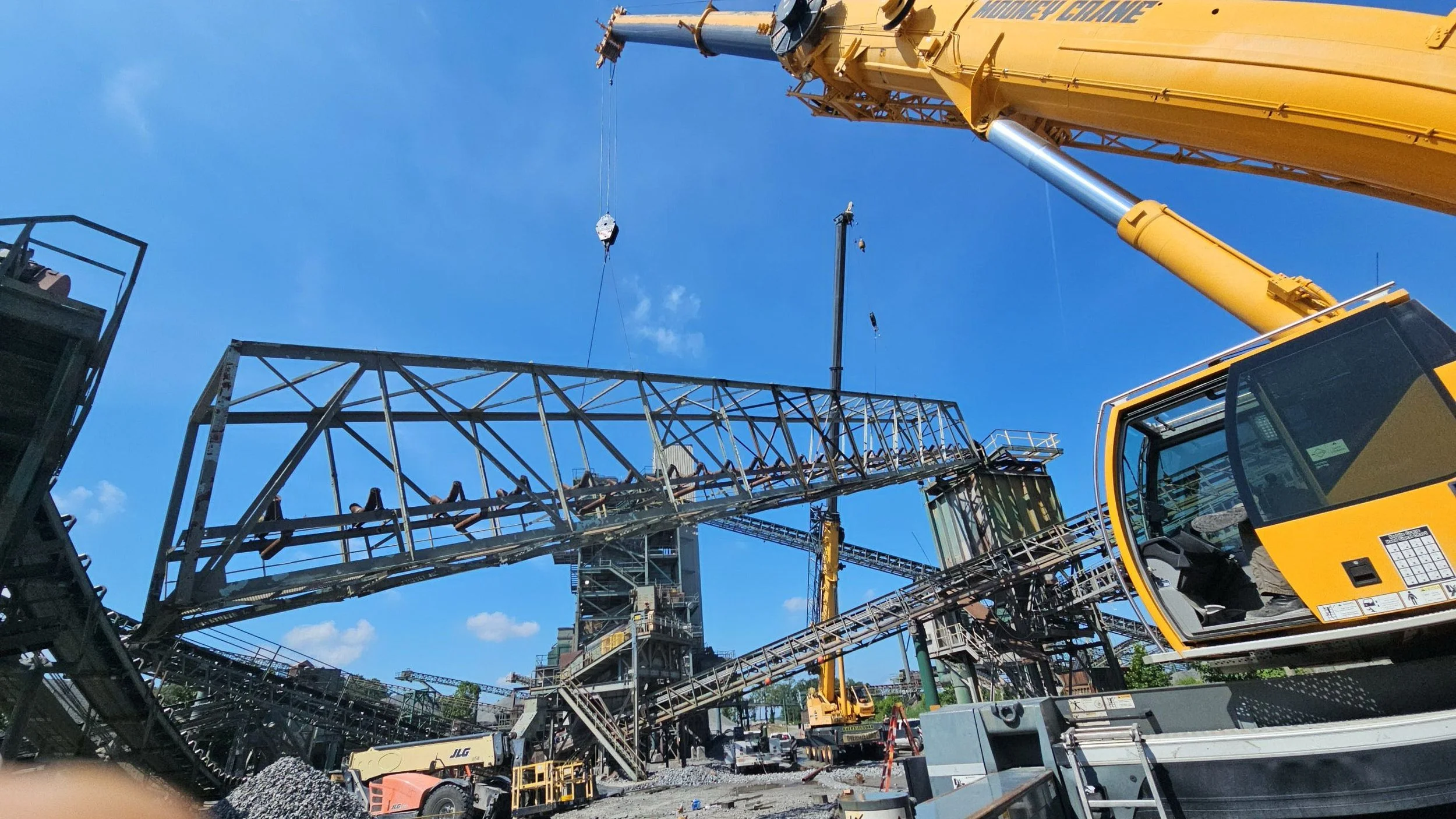 Construction site with a large yellow crane lifting steel framework, blue sky overhead, and various construction equipment and structures in progress.