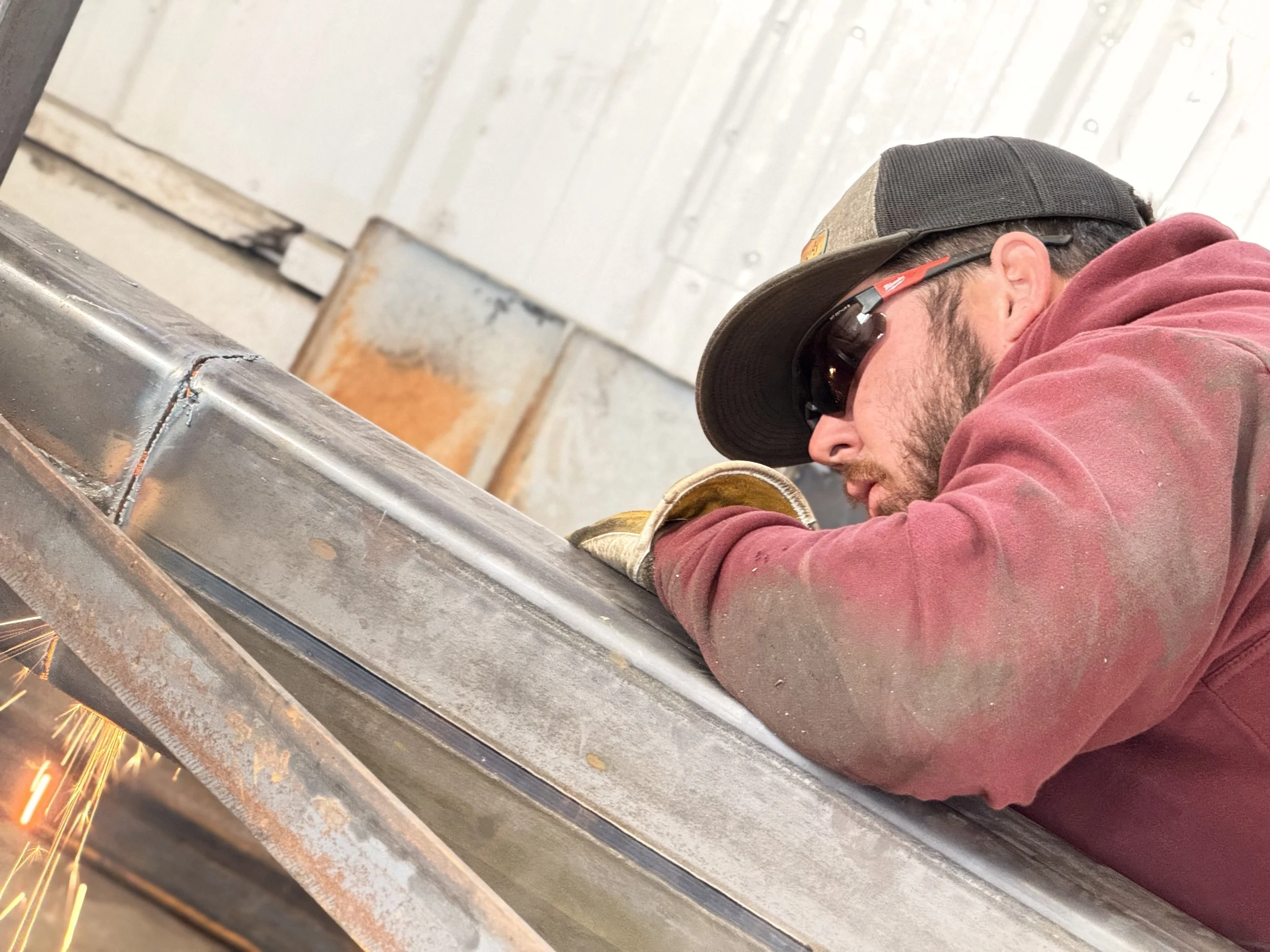 A man welding metal, wearing protective sunglasses, gloves, a hat, and a red jacket, sparks flying from the welding process.