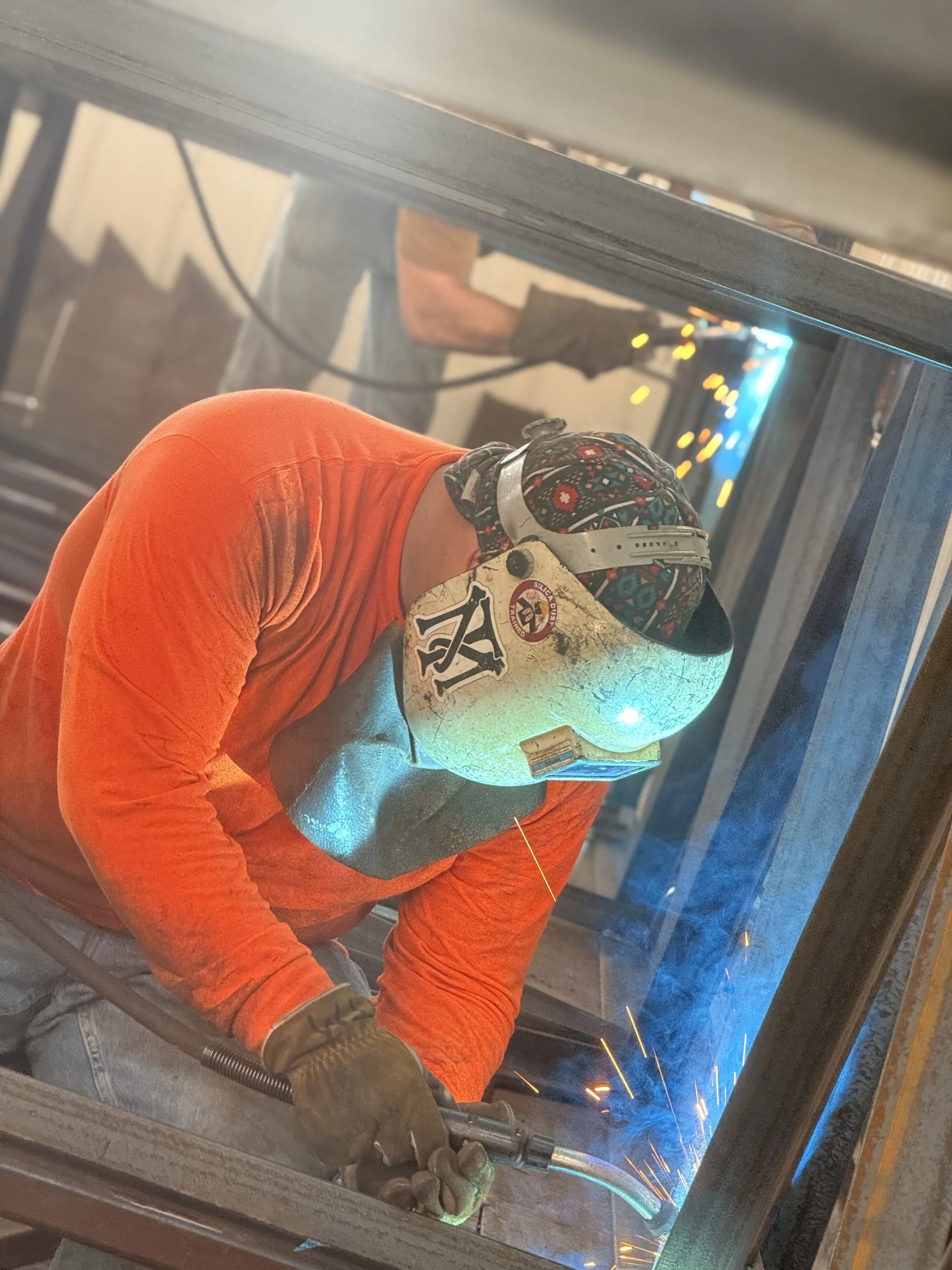 A worker welding metal inside a workshop, wearing a protective welding helmet, orange long-sleeve shirt, gloves, and a patterned bandana, with sparks flying from the welding process.