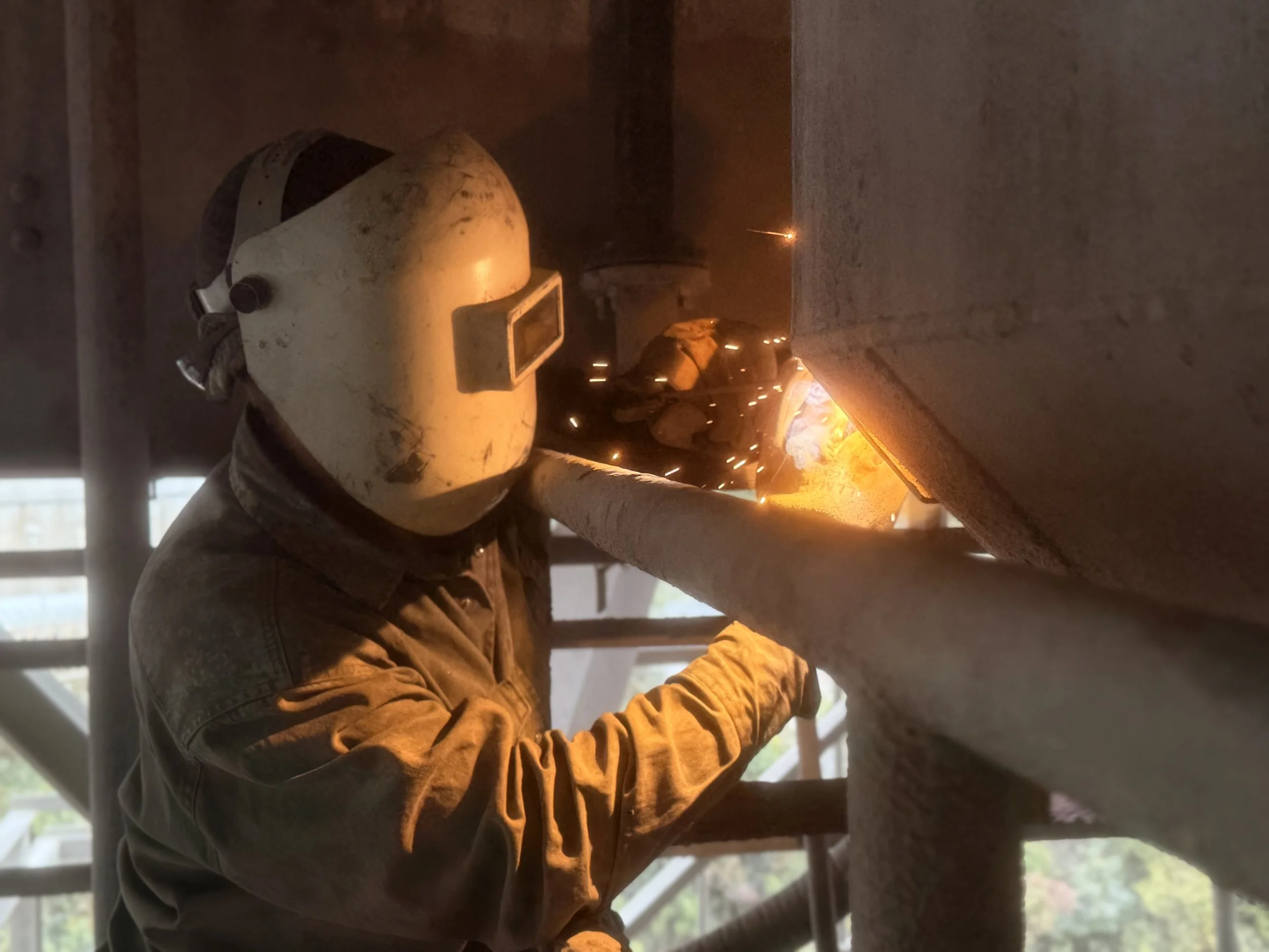Worker welding metal with protective helmet and gloves, sparks flying, in an industrial setting.