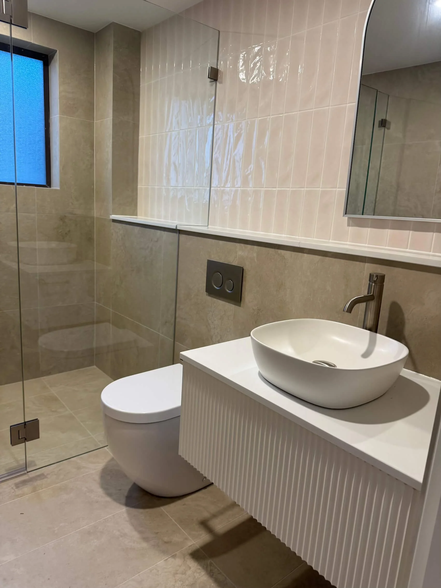 Modern bathroom with a wall-mounted sink in a vessel bowl style, a wall-mounted toilet, a large mirror, beige tiled walls and floor, and a glass shower enclosure with a window.