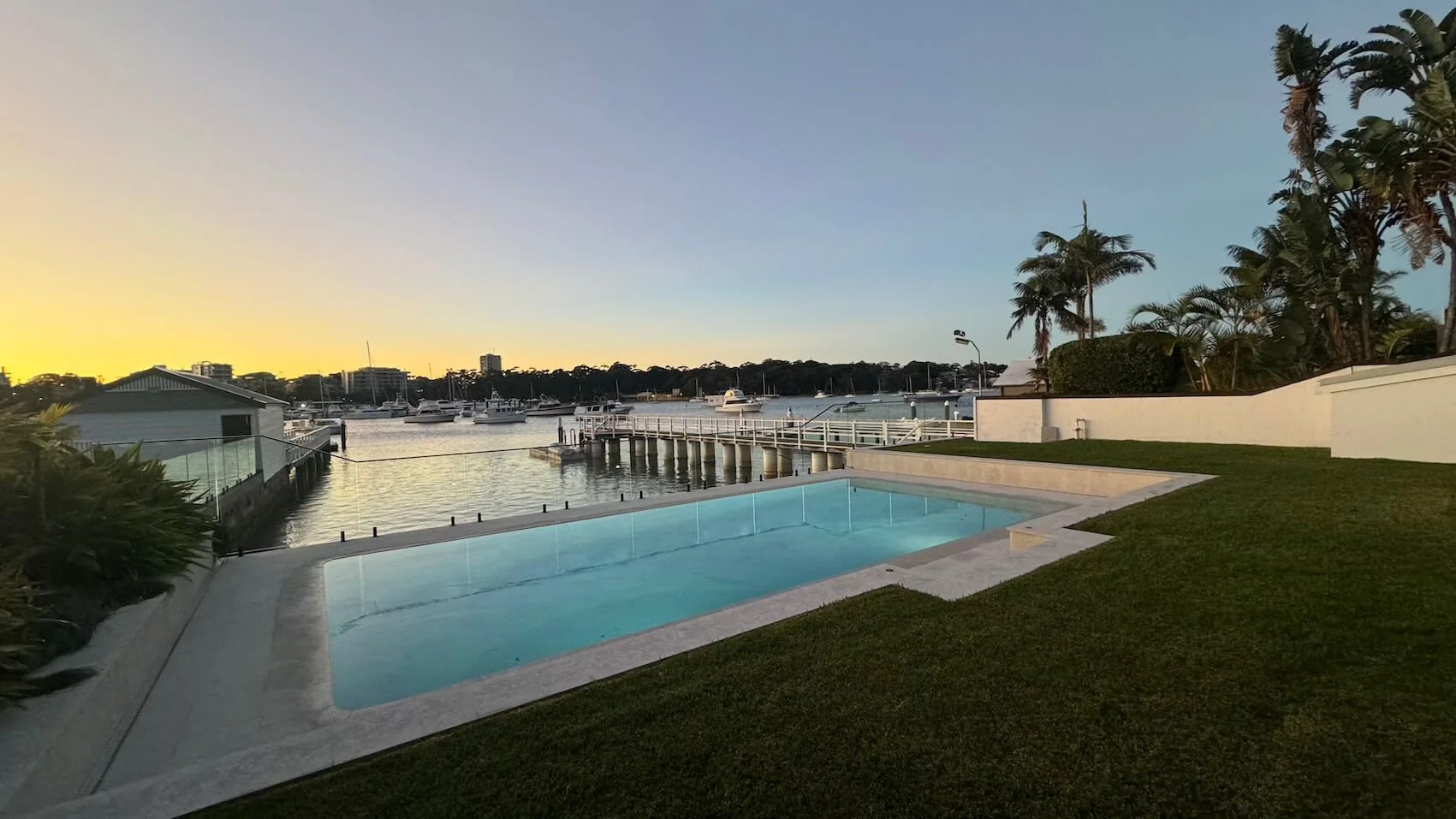 A backyard with an in-ground rectangular swimming pool, a grassy lawn, palm trees, and a waterway with boats and a dock during sunset or sunrise.