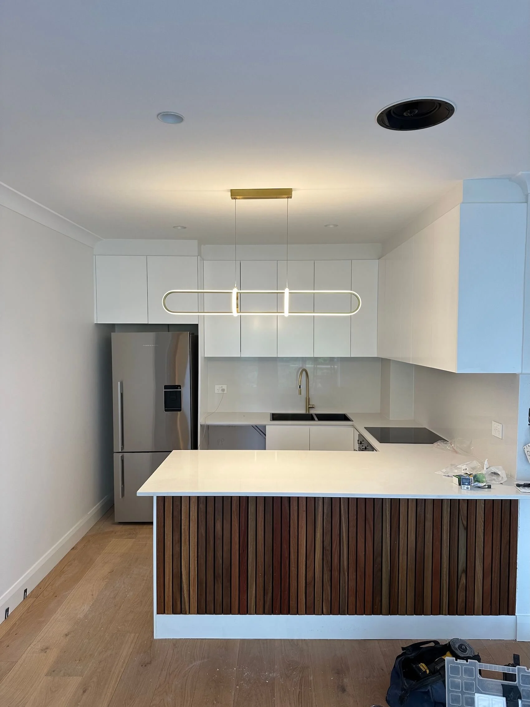 Modern kitchen with white cabinets, a stainless steel refrigerator, and a white island with a wooden front. Minimalist lighting fixture hangs over the island. The kitchen has a black sink and cooktop, with a brass faucet.