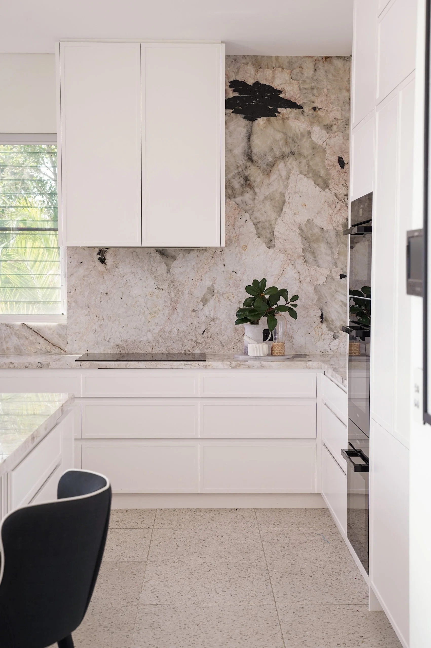 Modern kitchen with white cabinetry, a marble wall, a potted plant on the counter, and a window with green foliage outside.