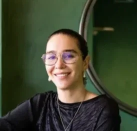 A woman with glasses smiling, with a round mirror and green wall in the background.