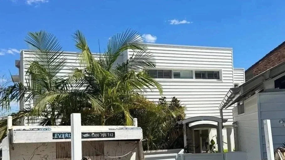 Modern white house with horizontal siding, surrounded by lush palm trees, under a bright blue sky.