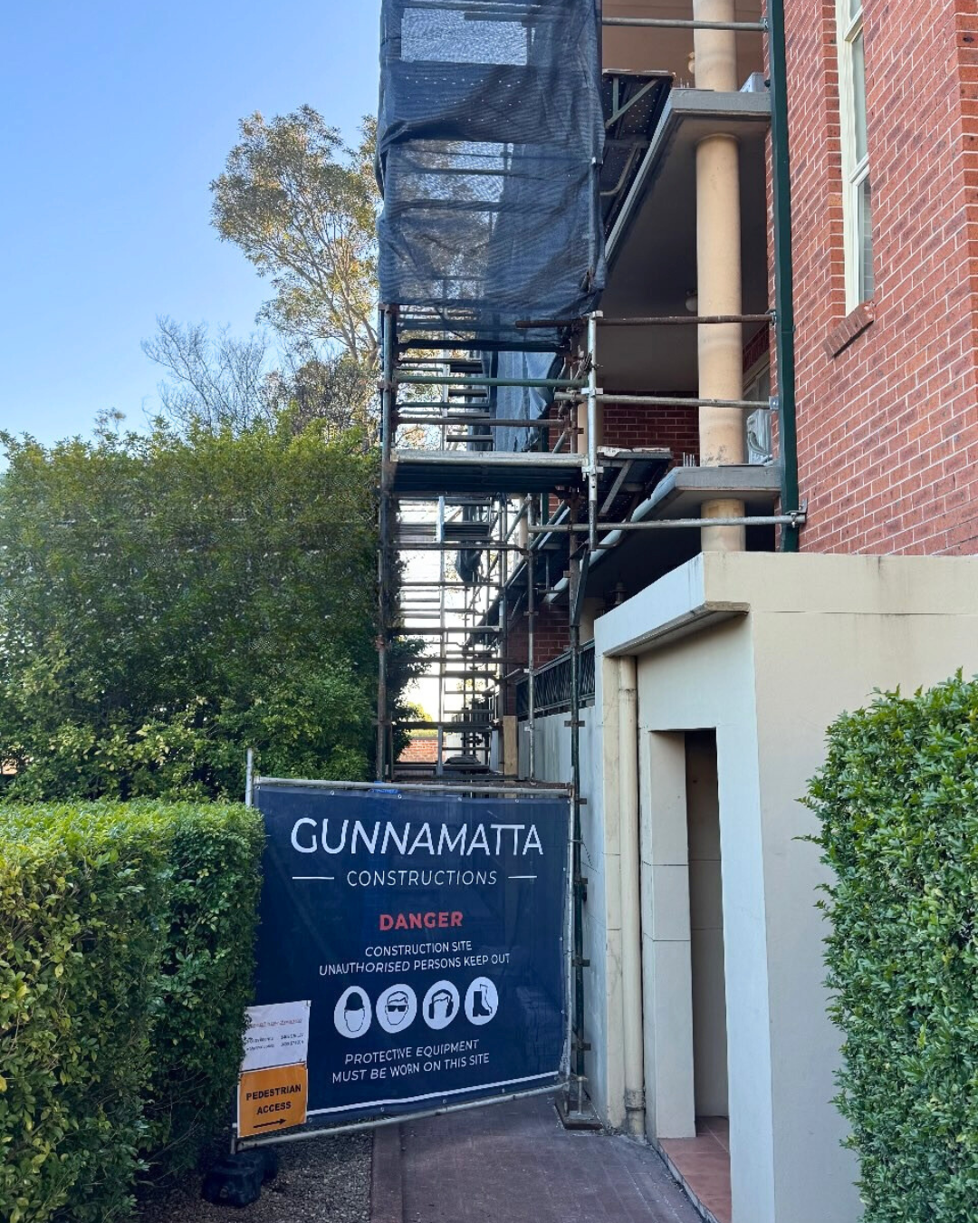 Construction site with scaffolding and safety sign reading 'GUNNAMATTA CONSTRUCTIONS' and warning about danger and protective equipment requirements.