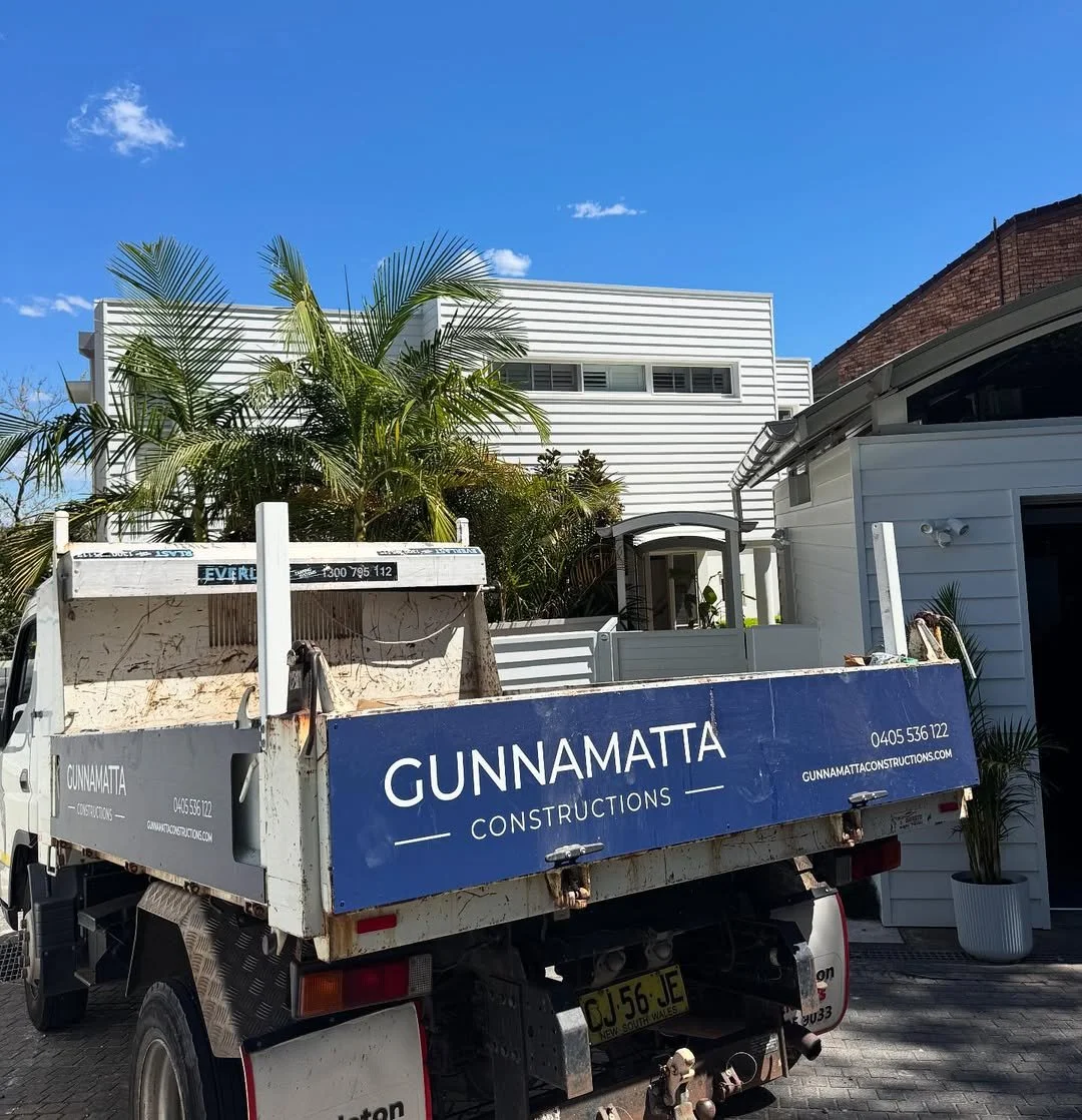Construction truck parked in front of a modern white house with large windows and palm trees under a blue sky.