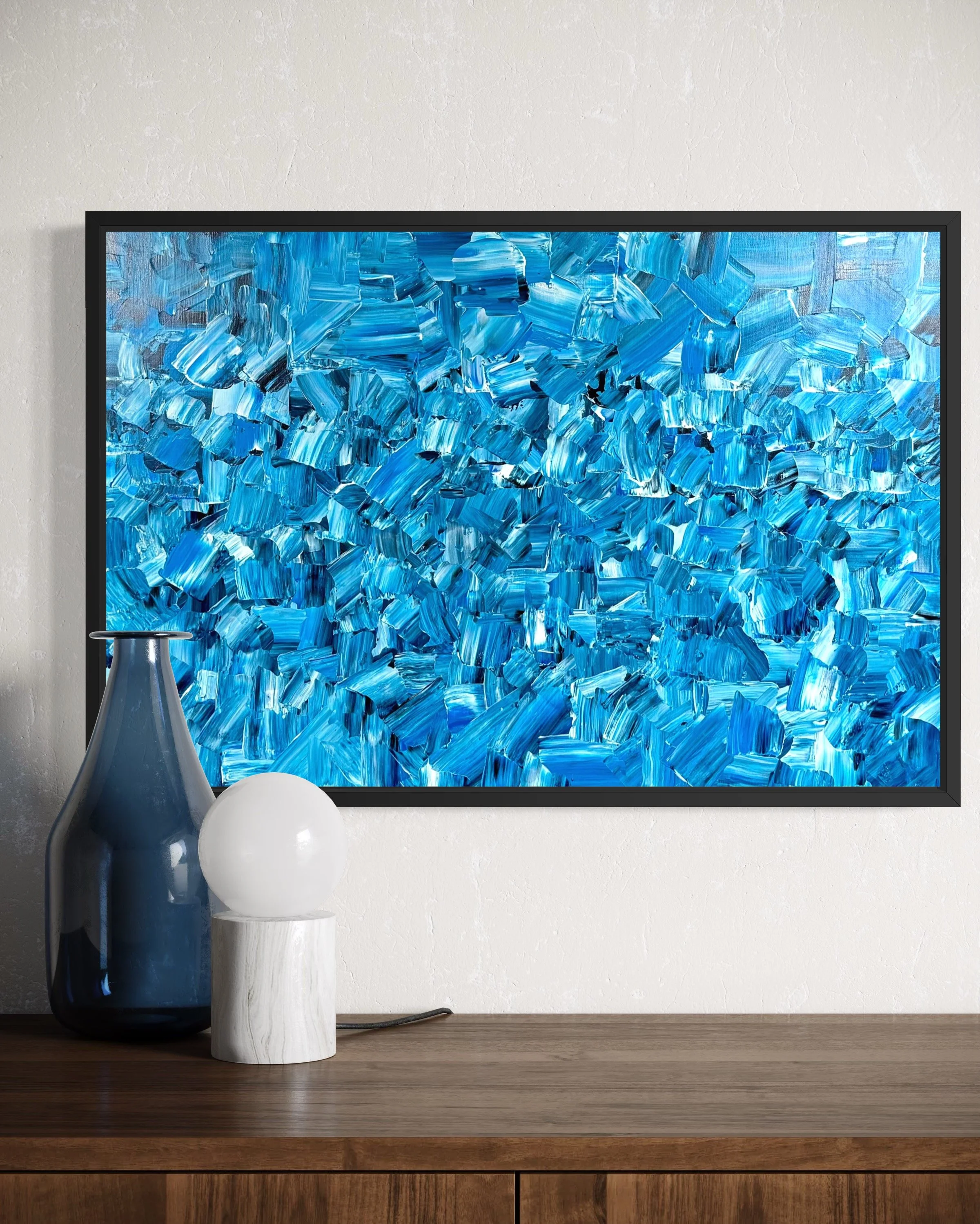 Abstract blue painting hanging on a white wall with a wooden surface below, decorated with a dark blue vase and a white spherical lamp.