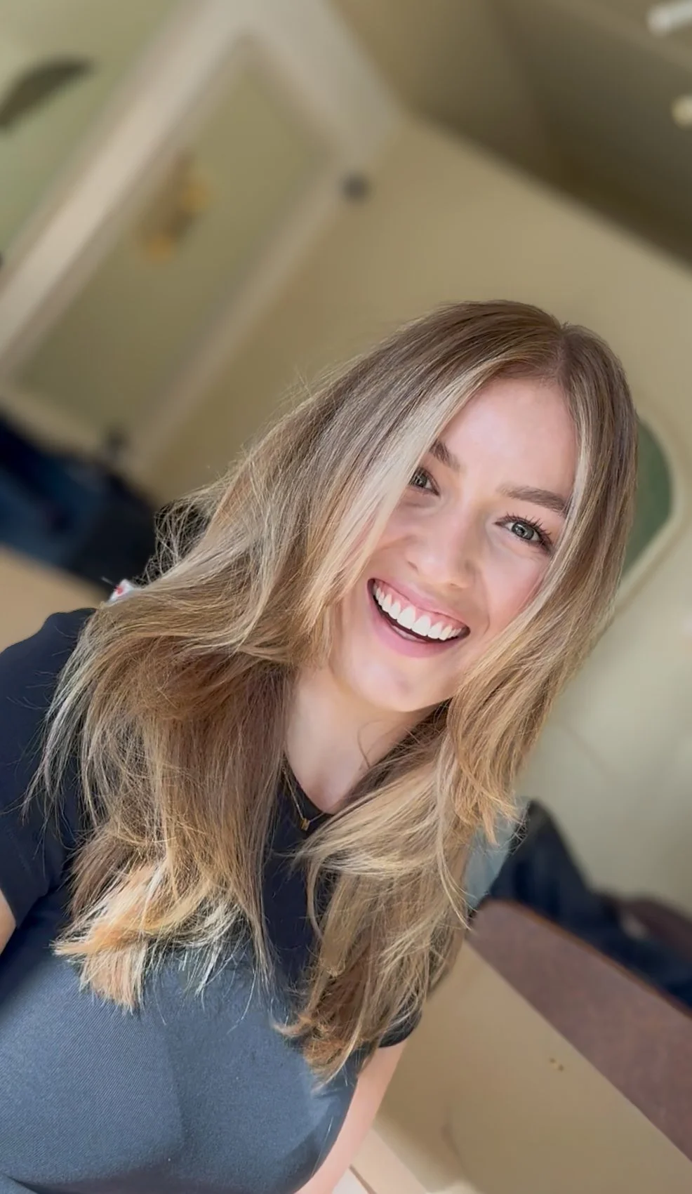A young woman with long, wavy blonde hair smiling brightly, wearing a black top, indoors with a blurred background.