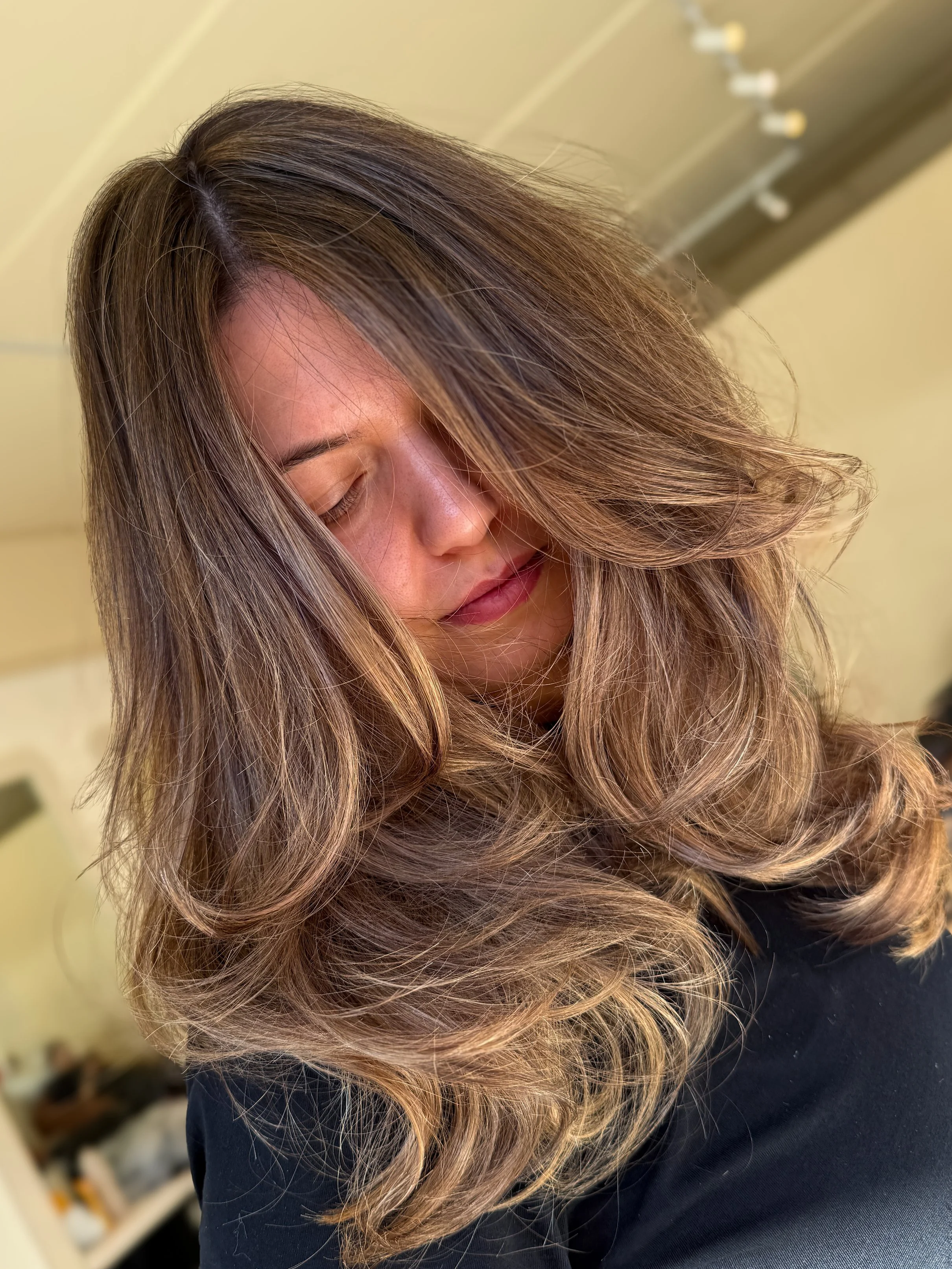 A woman with long, wavy brown hair with blonde highlights, looking downward with her eyes closed, in a hair salon.