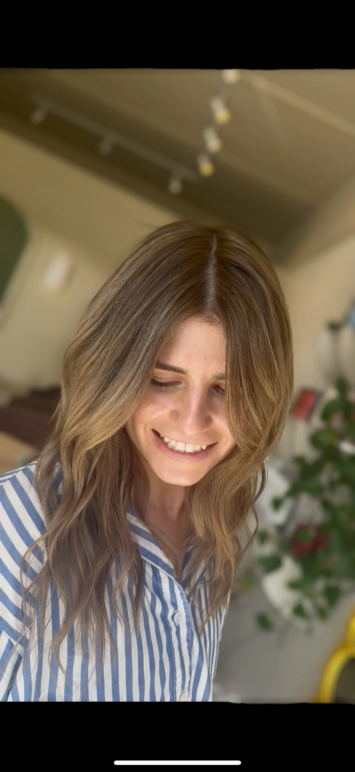 A young woman with long, wavy brown hair smiling in a striped shirt, indoors near a plant.