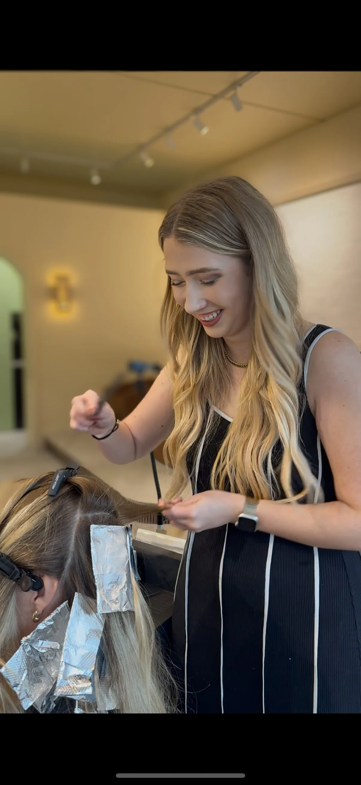 A woman with long wavy blonde hair getting her hair colored at a salon, with foil-wrapped hair sections, a stylist smiling and working on her hair.