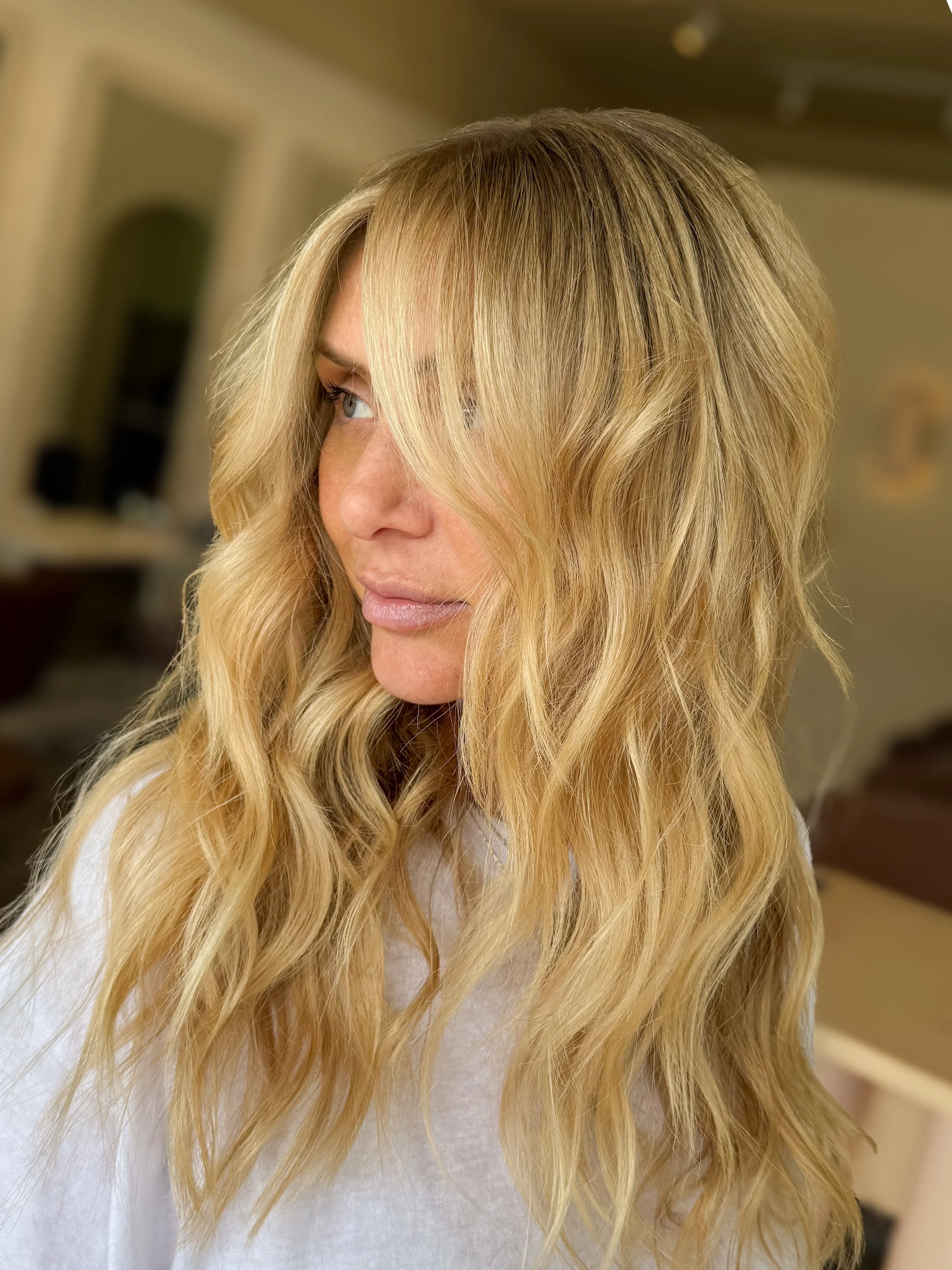 A woman with long, wavy blonde hair looking to the side, indoors with a blurred background.