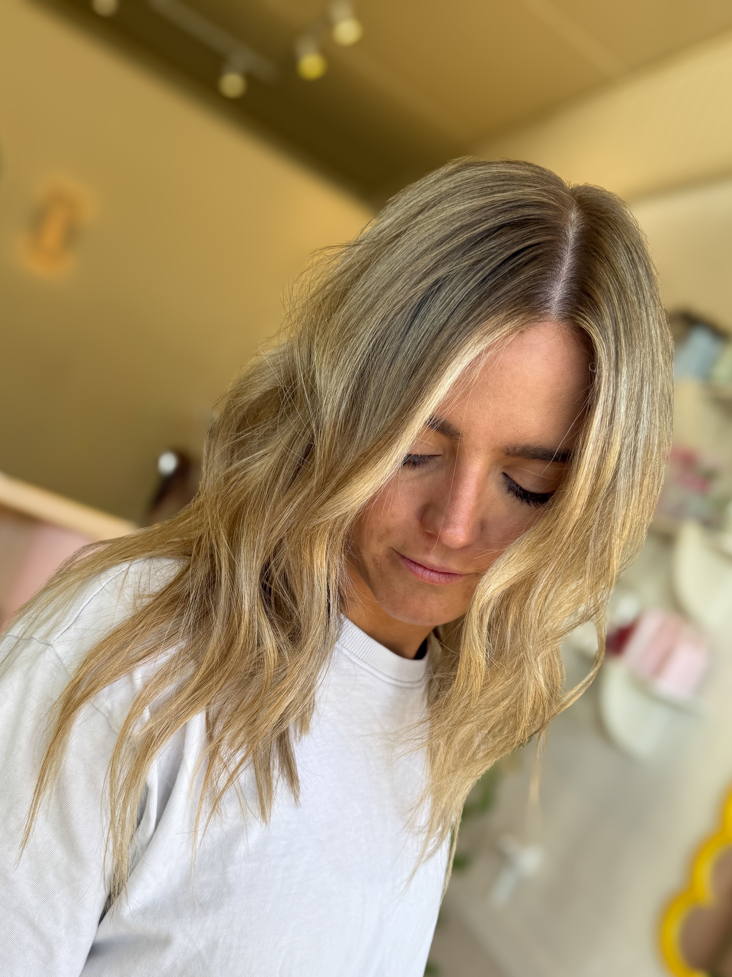 A woman with blonde, wavy hair wearing a white shirt, looking down with eyes closed in an indoor setting.