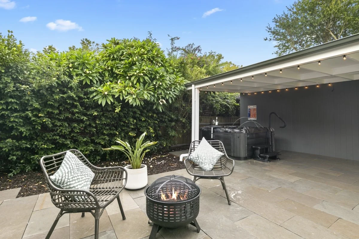 Patio area with two metal chairs, a potted plant, a fire pit, and a hot tub under a covered area with string lights, surrounded by greenery and trees.