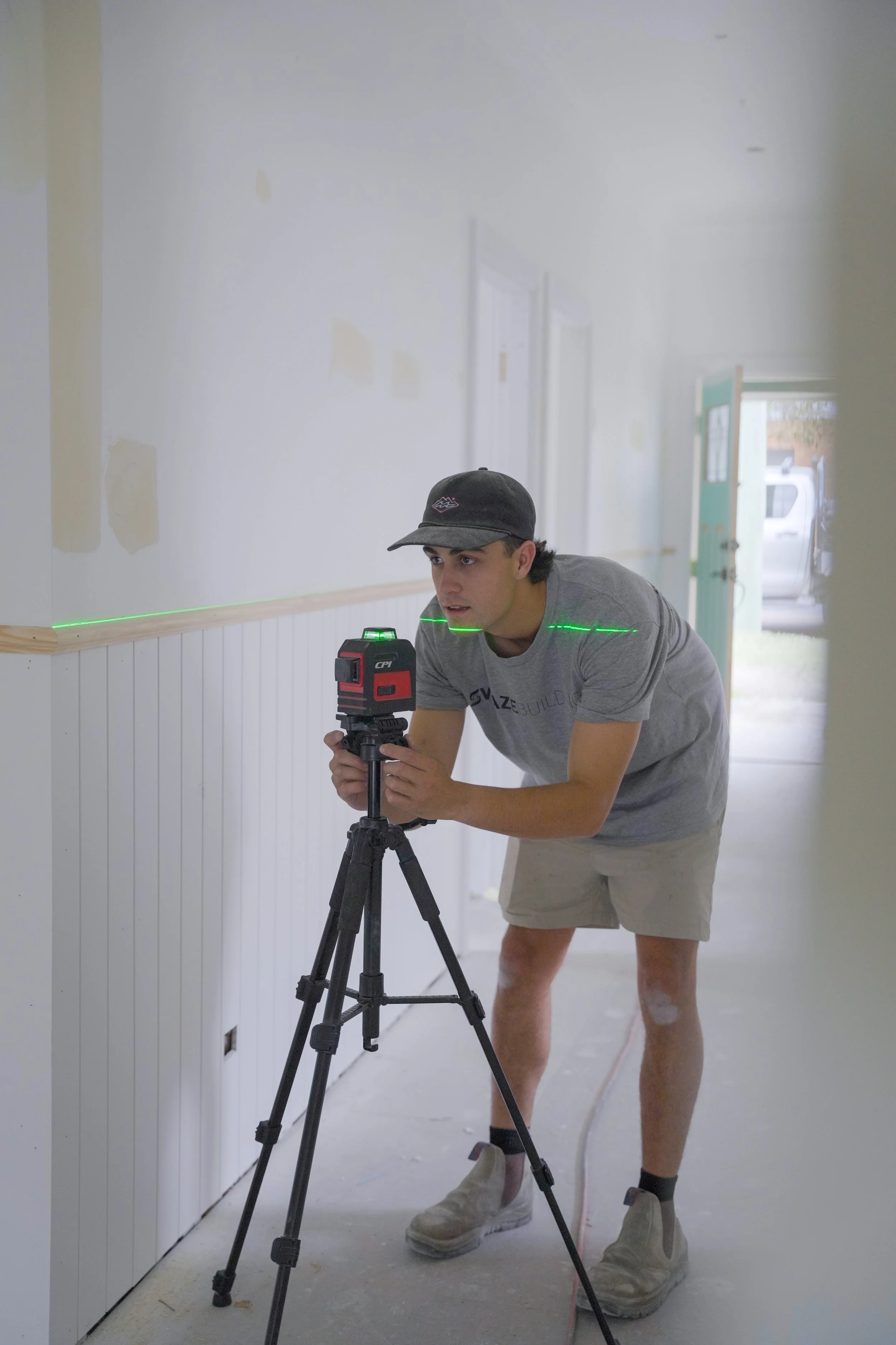 A man wearing a gray T-shirt, beige shorts, and a black cap is using a laser level on a tripod to measure or align a wall in a room under renovation, with visible patches of drywall and an open door leading outside.