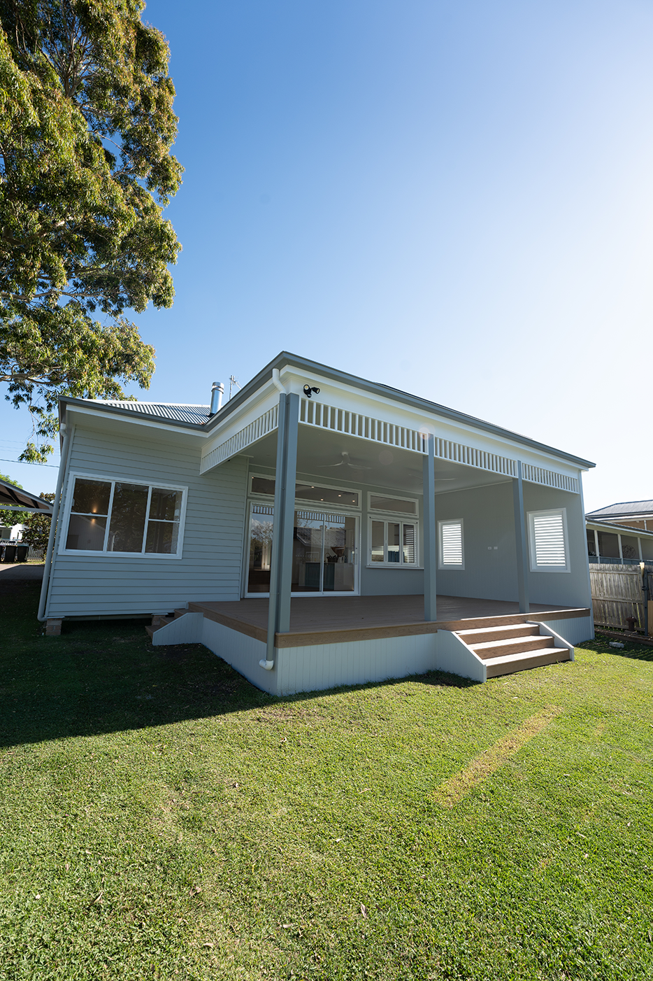 Back view of a modern white house with a patio, steps leading up to it, surrounded by a green lawn and a large tree on the left side, under a clear blue sky.