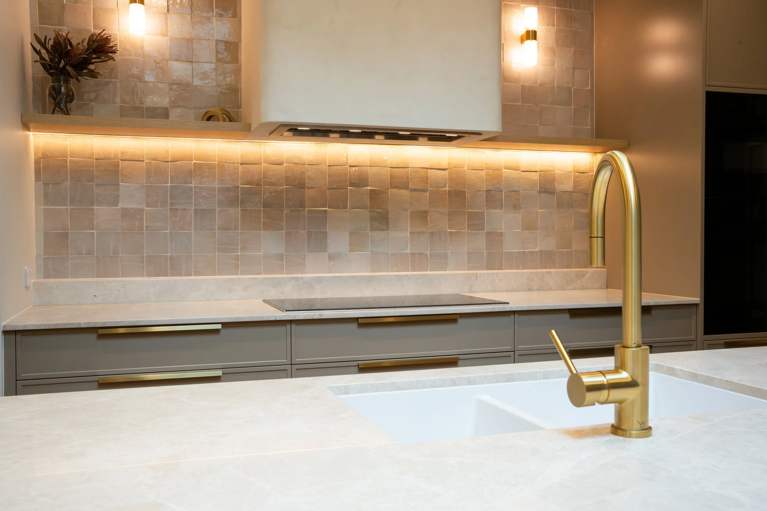 Modern kitchen with beige countertops, gold faucet, tiled beige backsplash, and a small vase with flowers on a wooden shelf