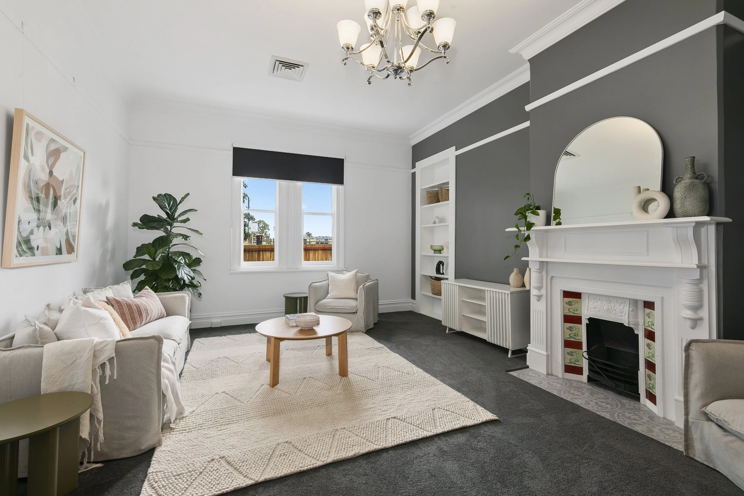 Living room with white walls, gray accent wall, white fireplace with decorative tiles, round mirror, built-in shelves, beige sofa with pillows, armchair, black window shade, chandelier, large green plant, natural-fiber rug, dark carpet