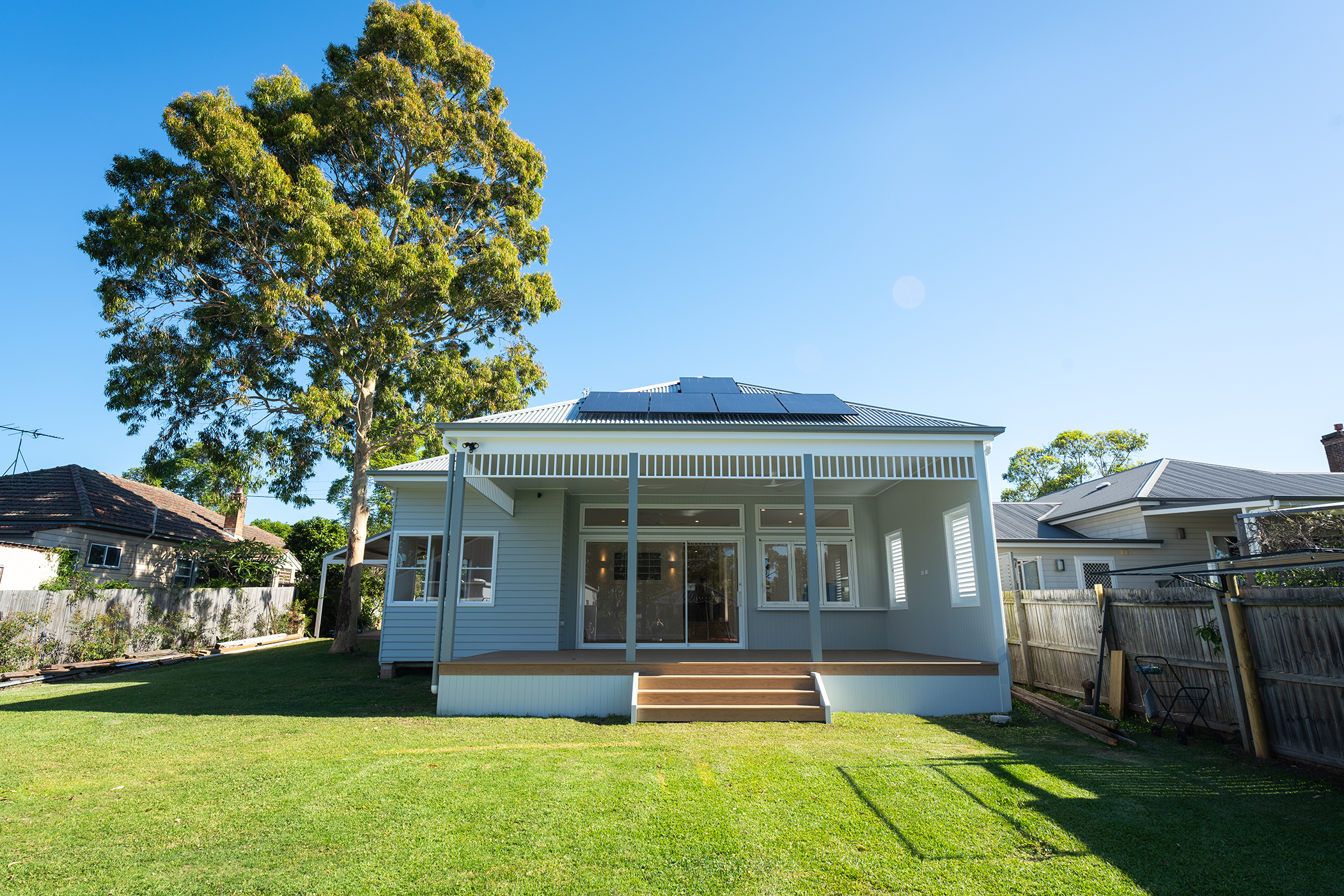 Back view of a modern two-story house with solar panels on the roof, a screened porch, wooden stairs leading to the yard, surrounded by a wooden fence, with a large tree on the left and neighboring houses visible.