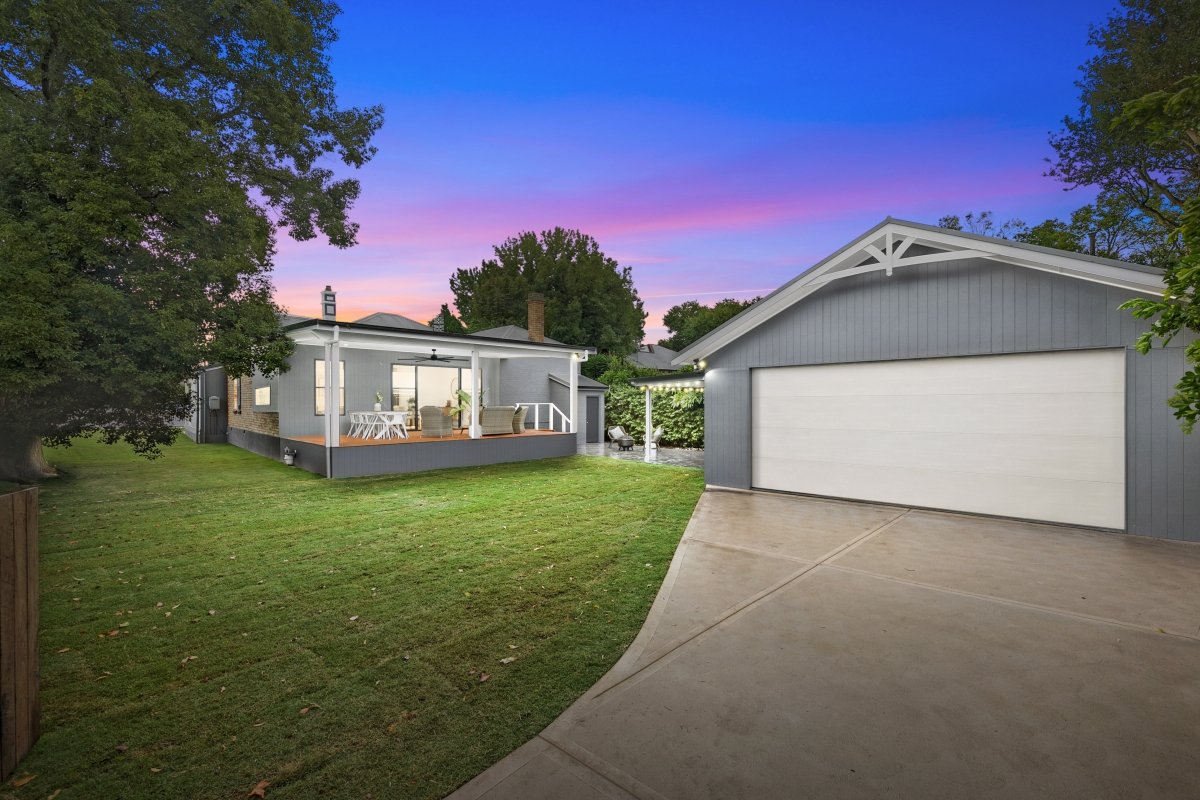 The image shows a backyard at dusk with a gray house featuring a front porch with outdoor furniture. There is a garage on the right side, and the sky displays a colorful sunset with shades of purple and pink.