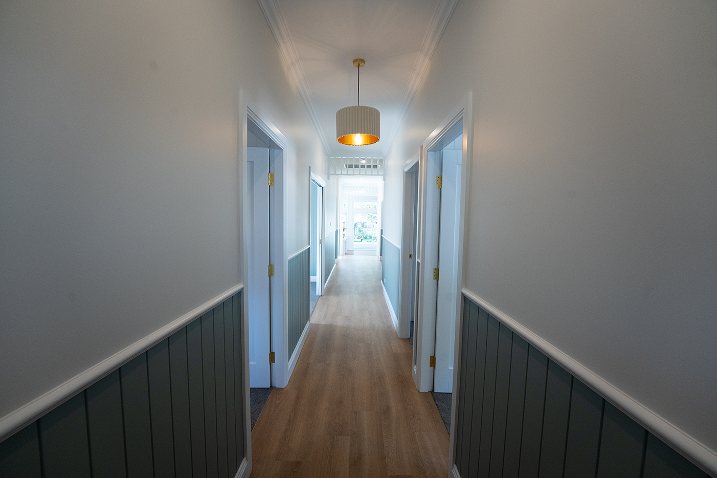A hallway with white walls, dark green wainscoting, light wood flooring, and a ceiling light fixture, leading to a bright door at the end.
