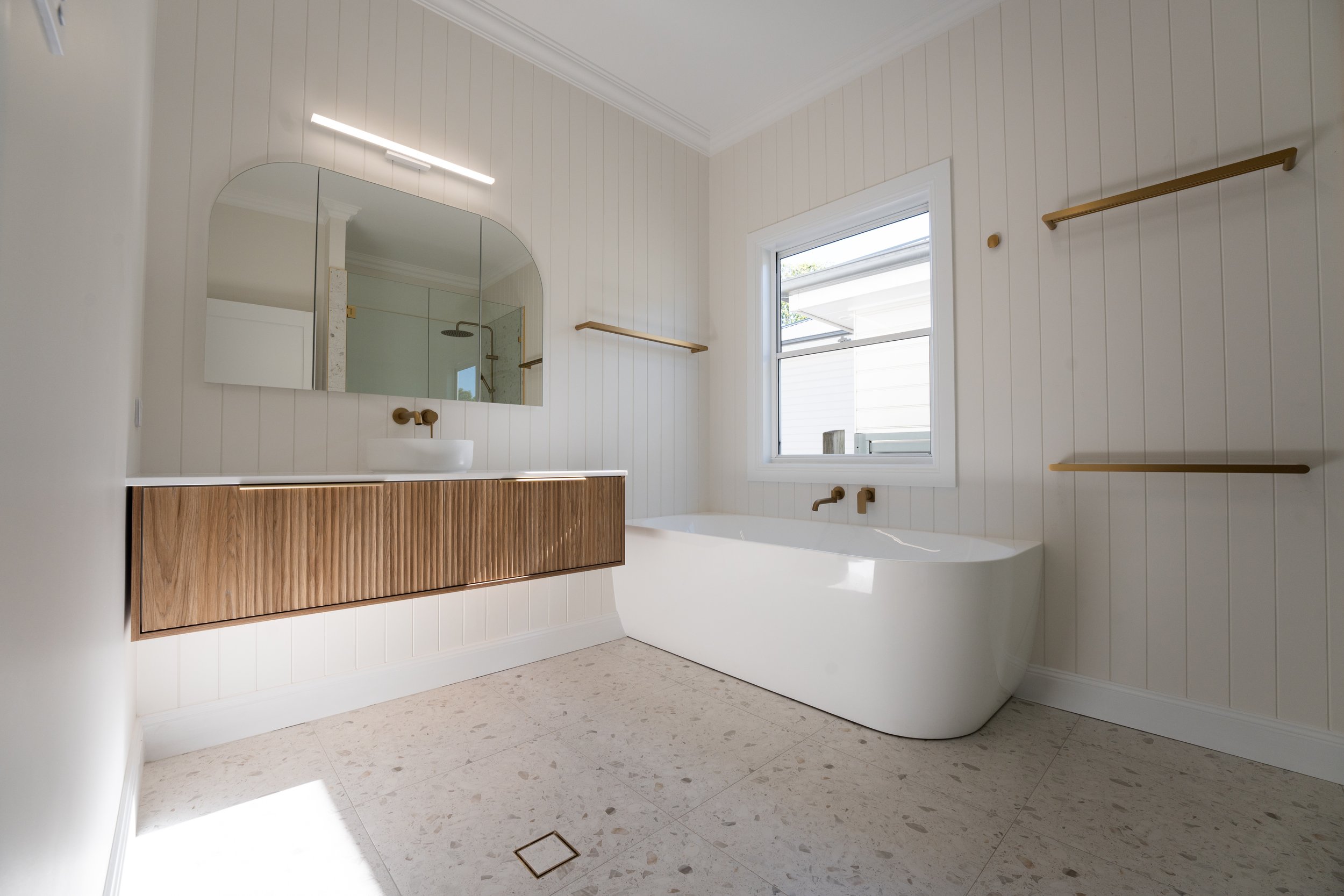 Modern bathroom with a freestanding bathtub, wooden vanity with white vessel sink, large wall mirror, window, and minimal gold-colored shelves.