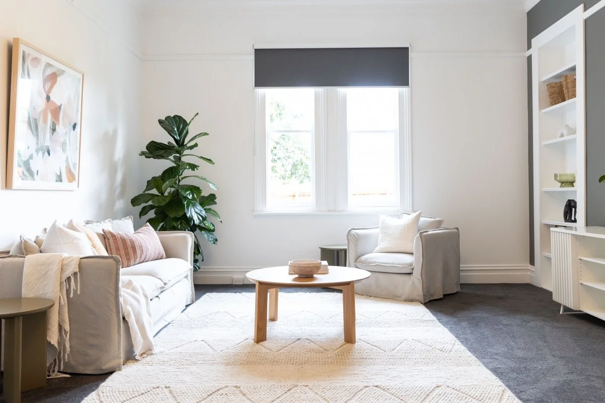 Bright living room with white walls, a large beige sofa with pillows, a white armchair, a round wooden coffee table, a large leafy green plant, a window with a dark gray blind, a white bookshelf with decorative items, and a beige rug on a dark carpeted floor.
