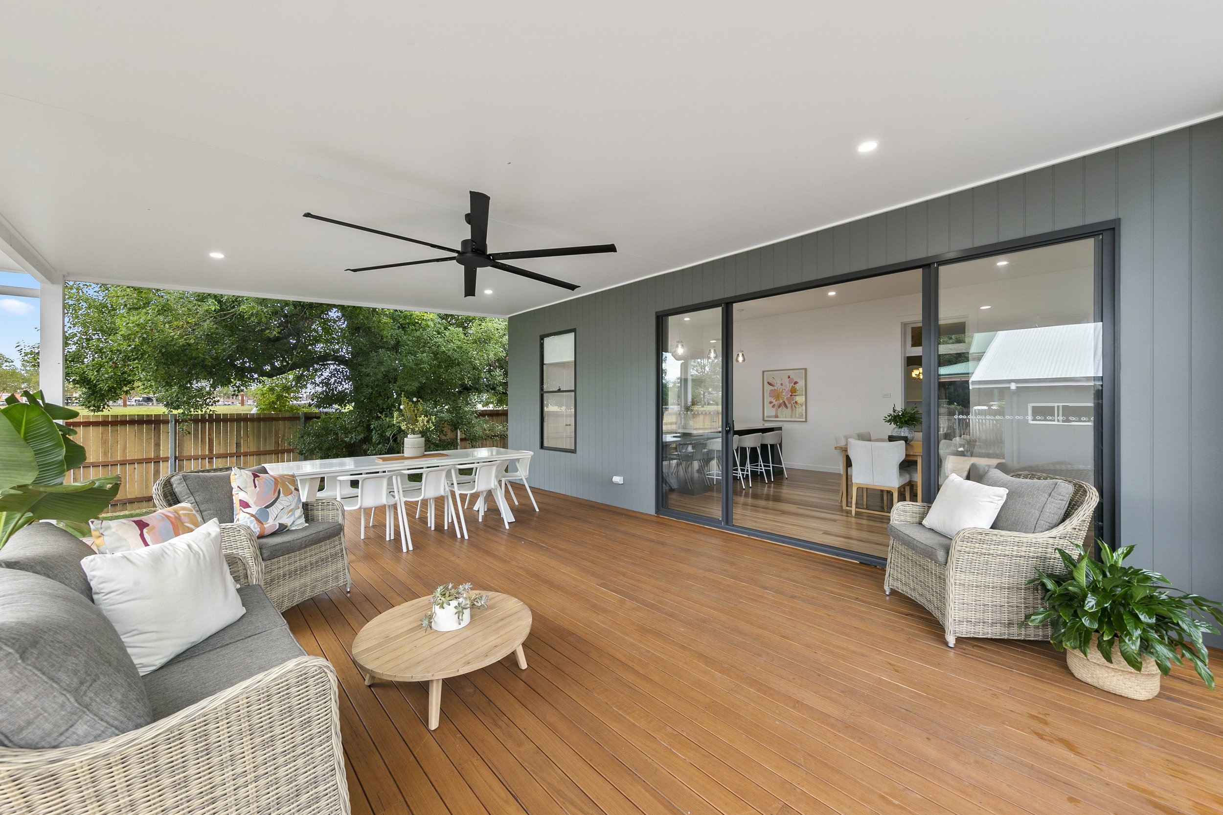 Spacious outdoor patio with wicker furniture, a gray sofa with pillows, a small wooden coffee table, a white dining table with white chairs, and potted plants. The area is covered with a white ceiling featuring a black ceiling fan, and the patio has wooden flooring. An interior room with wooden flooring and artwork can be seen through sliding glass doors.