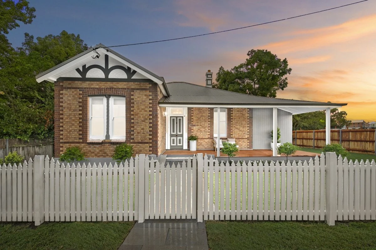 A charming brick house with a front porch, white picket fence, and small manicured shrubs, captured at sunset.