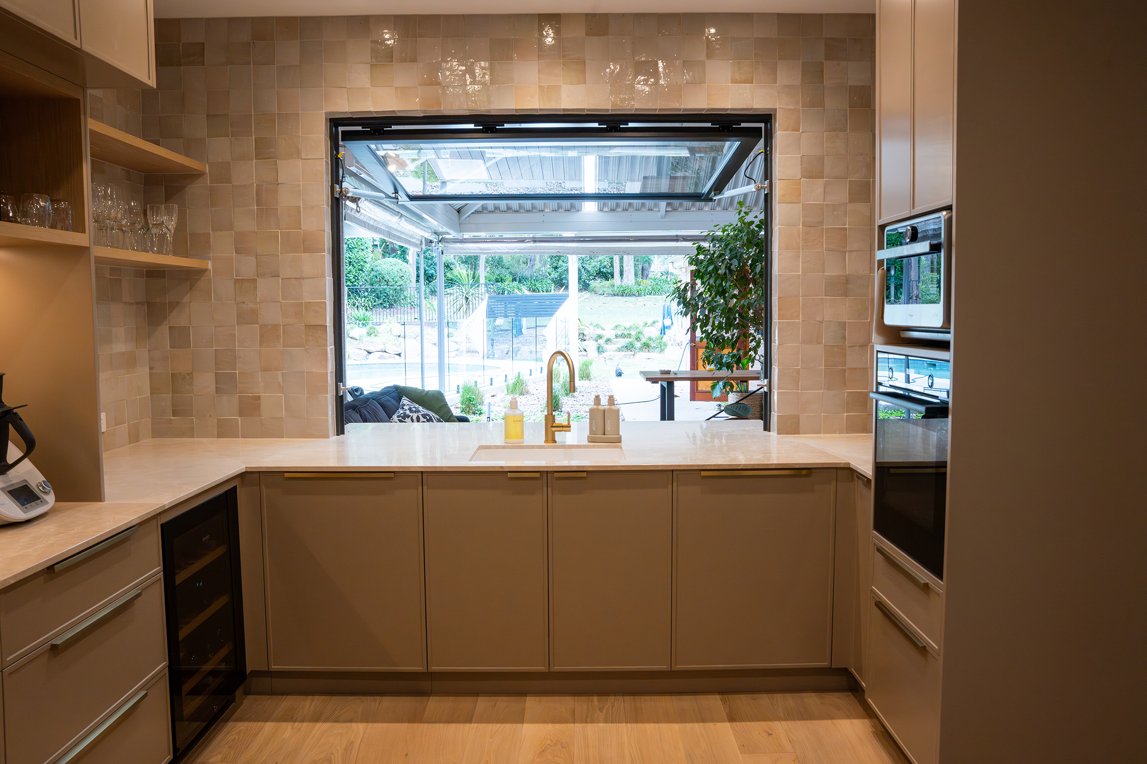 View of a modern kitchen with a large window looking into an outdoor patio and backyard, beige cabinets, a built-in wine cooler, and a marble countertop.