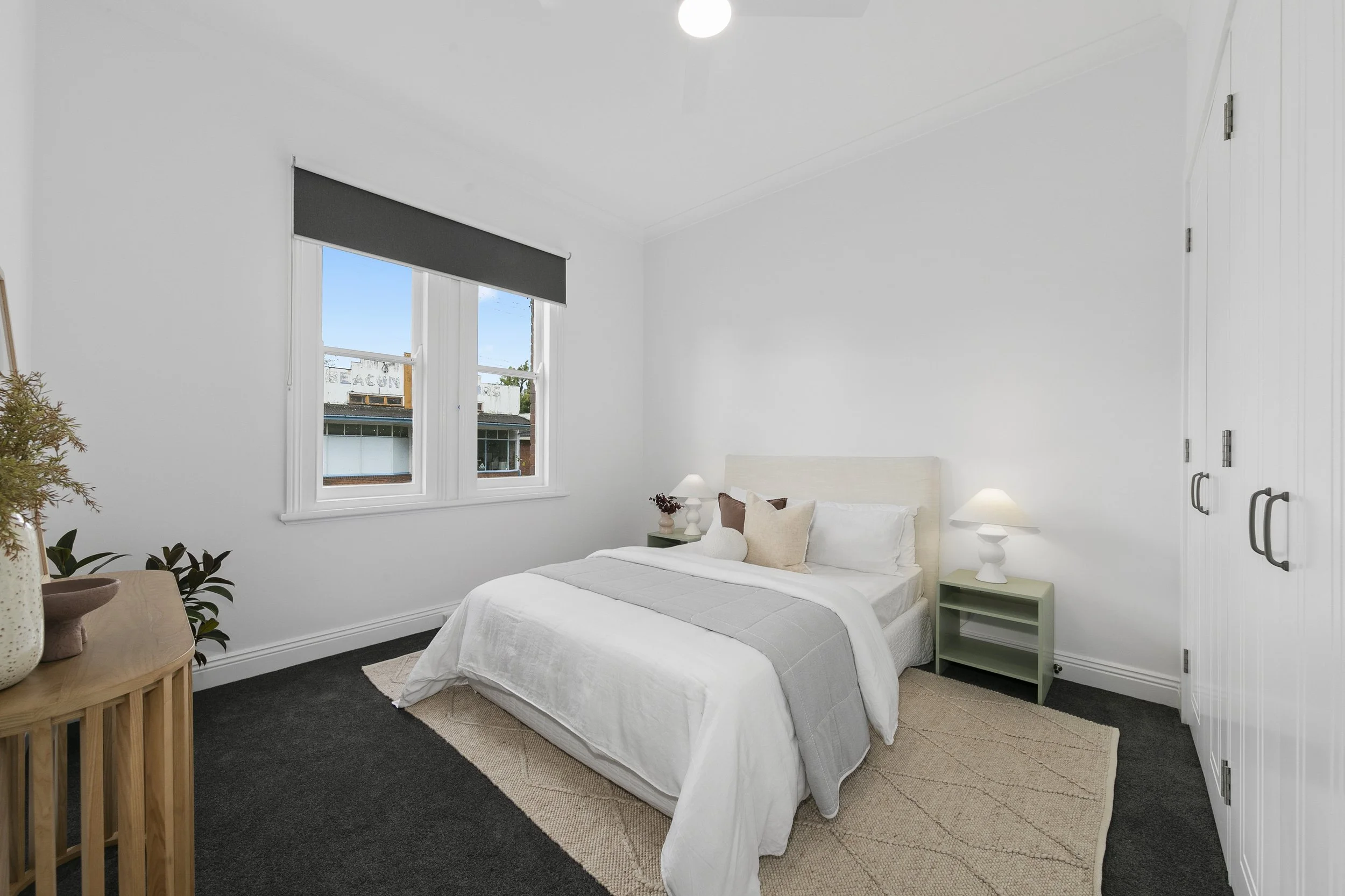 A bright bedroom with white walls, a bed with white and beige bedding, two nightstands with lamps, and a window with a view of buildings outside.