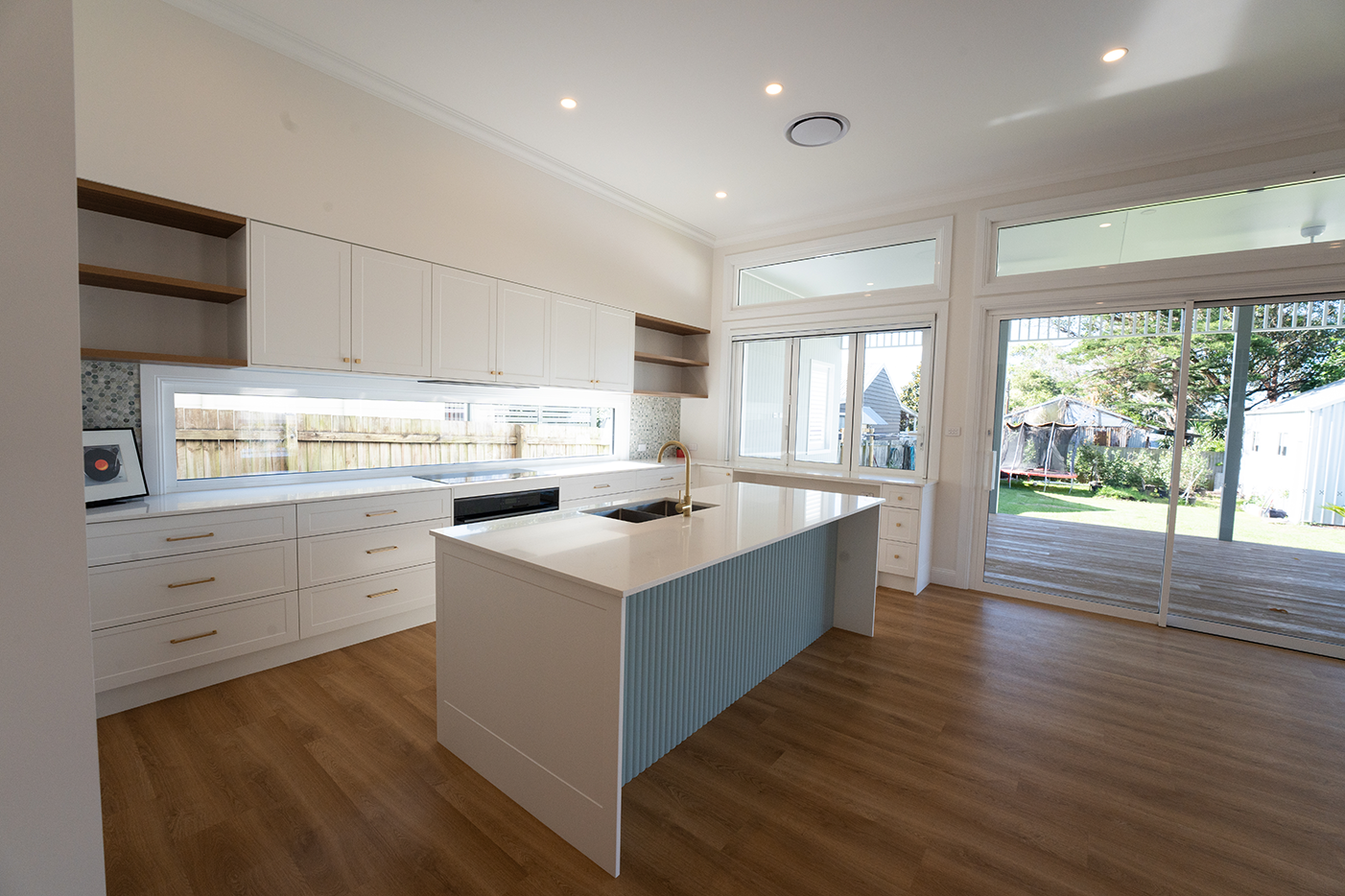 Modern kitchen with white cabinets, a kitchen island with a light blue panel, wooden flooring, large windows, and a sliding glass door leading to an outdoor deck and yard.