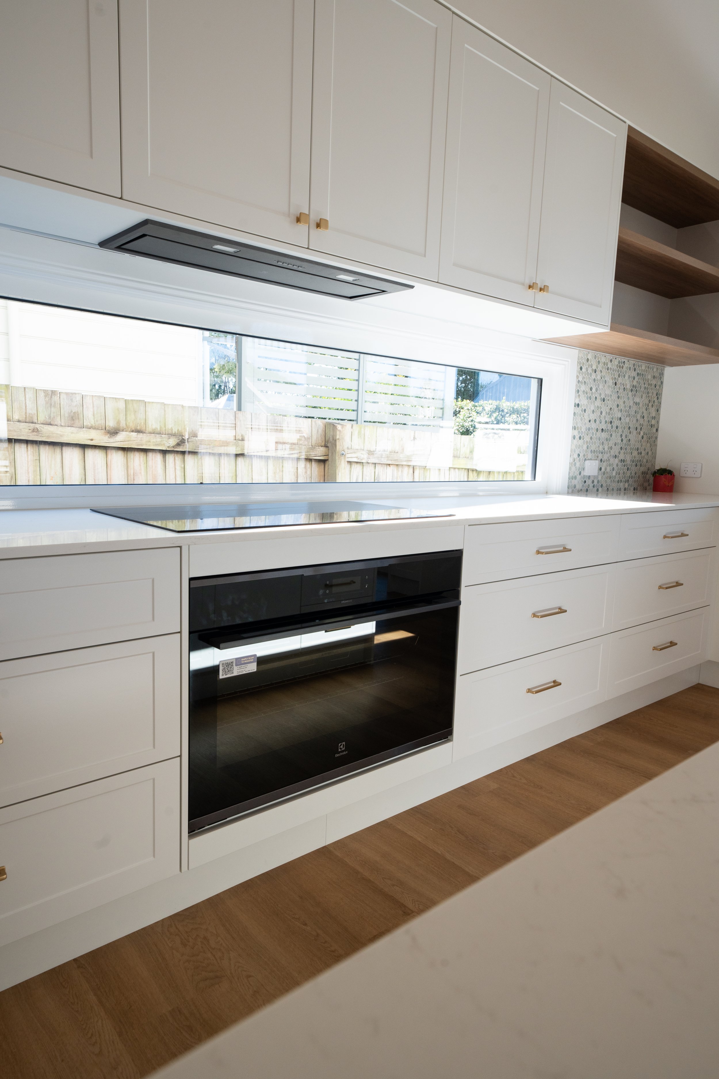 Modern kitchen with white cabinets, a black built-in oven, a white countertop, and a window overlooking a backyard with a wooden fence.