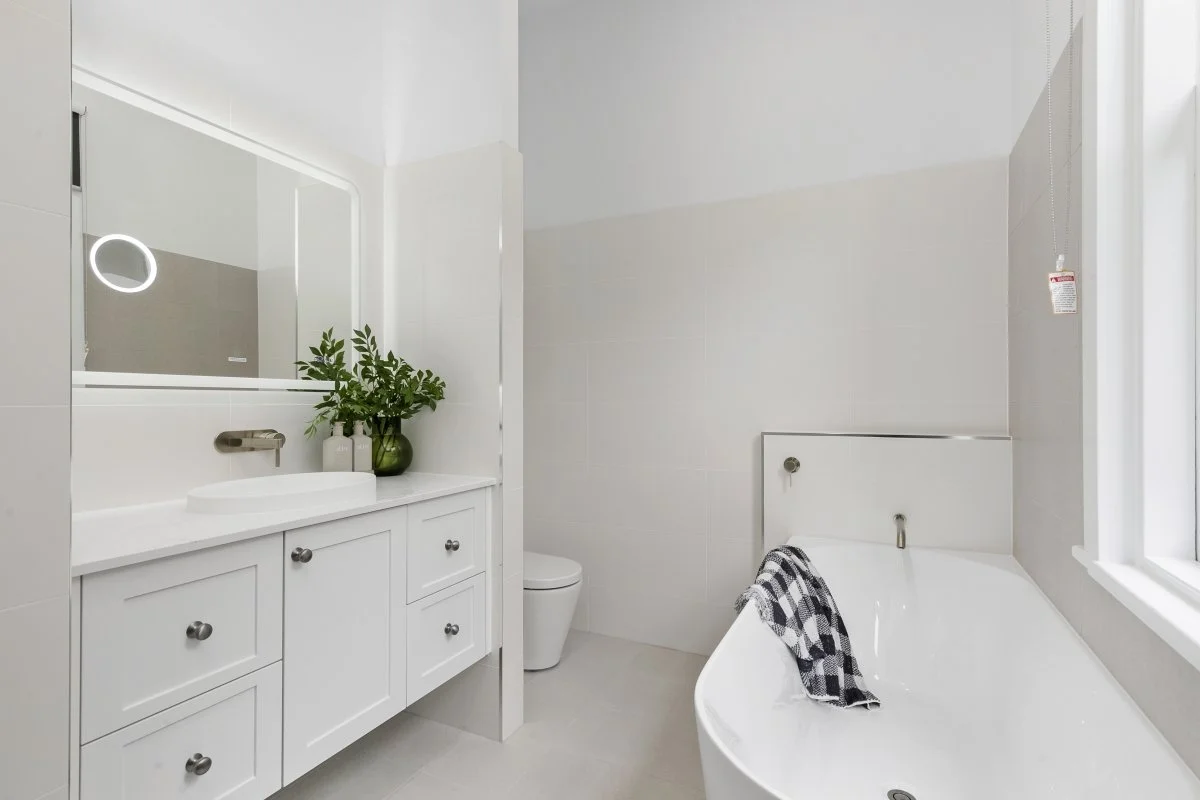 Modern bathroom with white vanity, large mirror, green plant, white sink, toilet, and bathtub with black and white towel.