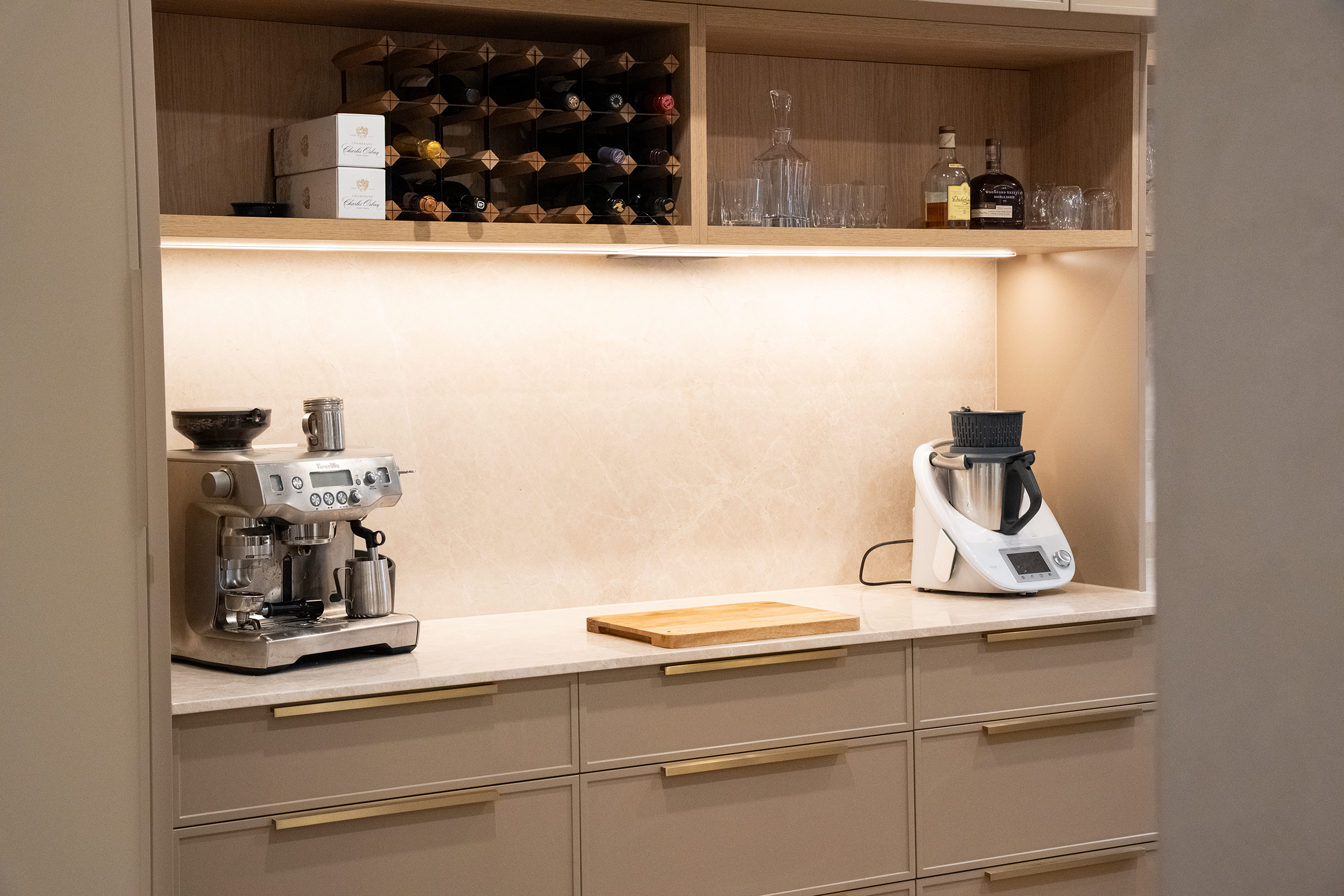A modern kitchen counter with a coffee machine, a cutting board, and an immersion blender, with open shelves above containing wine bottles, glasses, and liquor bottles.
