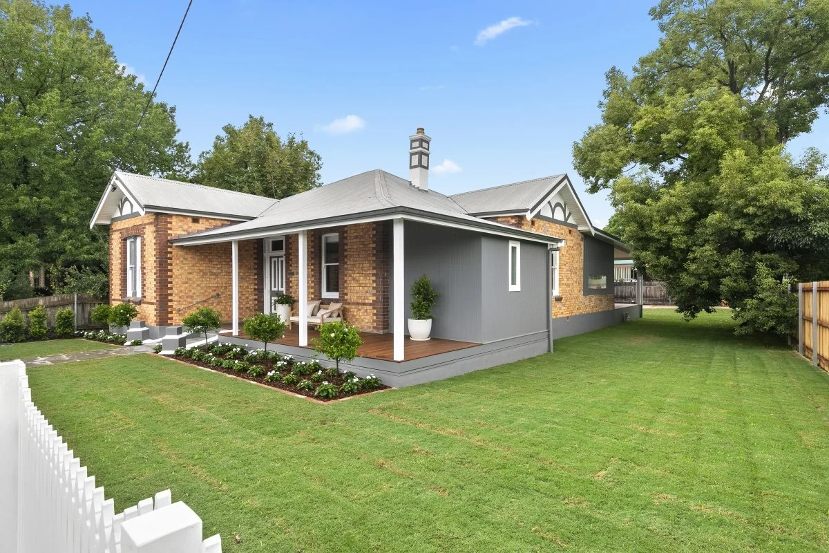 A house with a brick exterior, painted gray siding, a metal roof, and a small porch with chairs. The yard has a well-maintained lawn, flower bed, and trees with green leaves.