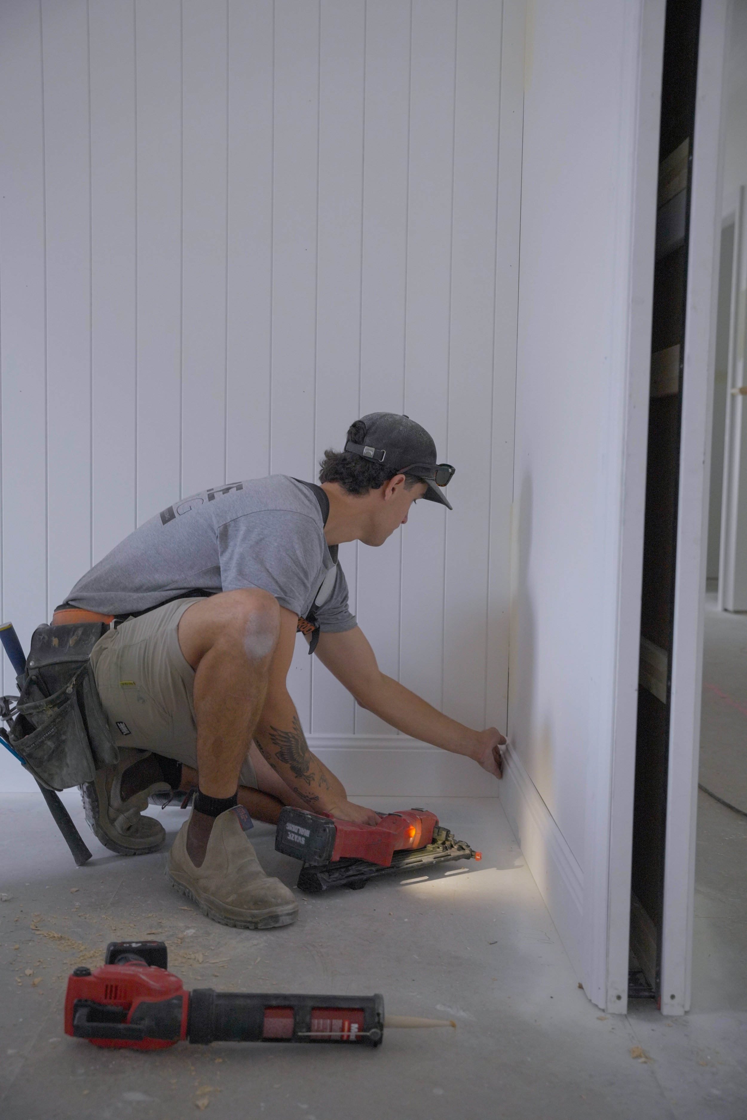 A man crouching down, working on baseboard molding in a room with white panel walls. He has a tattoo on his left leg, is wearing a gray cap, gray T-shirt, khaki shorts, and beige work boots. Power tools and sawdust are on the floor.